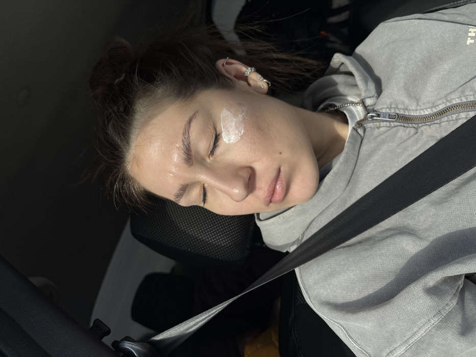 A female subject is depicted from the mid-chest upward, seated in what appears to be the front passenger seat of a vehicle, secured by a dark seatbelt across her torso. Her eyes are closed, and her head is tilted slightly to her right, with direct sunlight illuminating the left side of her face. A dollop of white cream or lotion is visible on her left cheekbone, below the outer corner of her left eye. She has dark hair pulled into a messy bun and wears a light grey zippered hoodie over a white undershirt with a thin silver chain visible at her neck. Her left ear features multiple piercings, including various studs and rings, one shaped like a flower. The vehicle's interior appears dark, with a black headrest directly behind her. No specific location within Ustinovo, Russia, or any other city, can be determined from the visual evidence presented in the image.