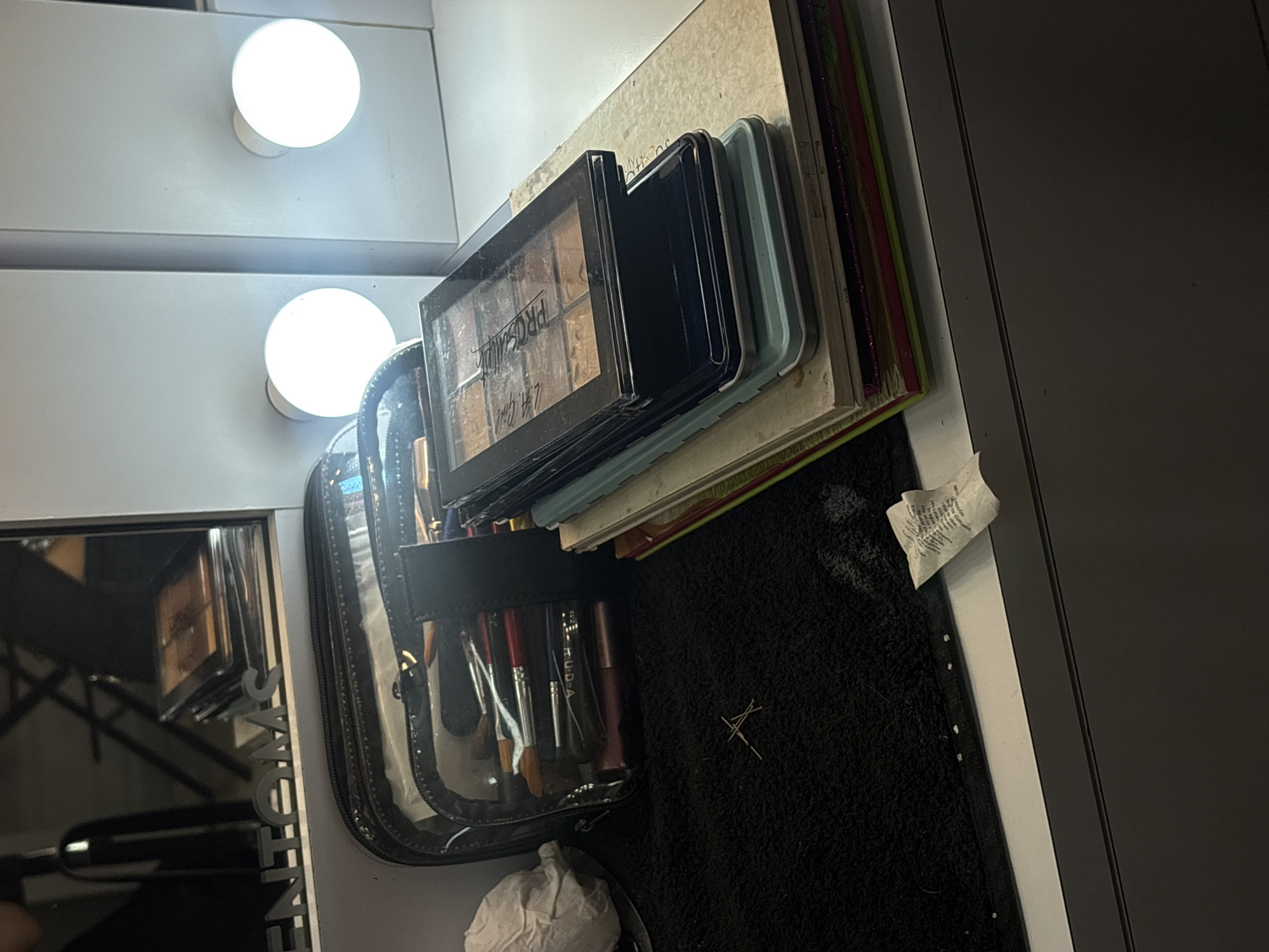The image depicts a vanity area in what appears to be a domestic setting. A lighted mirror is visible on the left, with two round light bulbs illuminated above. The vanity surface is cluttered with makeup products, including a palette labeled "ProSculpt," and various other palettes stacked together. A makeup brush holder, filled with an assortment of brushes and cosmetic items, stands next to the palettes. A black textured surface, possibly a vanity mat, lies beneath the makeup items. There is a crumpled tissue or paper near the brush holder. A dark grey wall is visible on the right side of the frame. The location is speculated to be in Mexico City, Mexico, but there is no data to confirm this information.