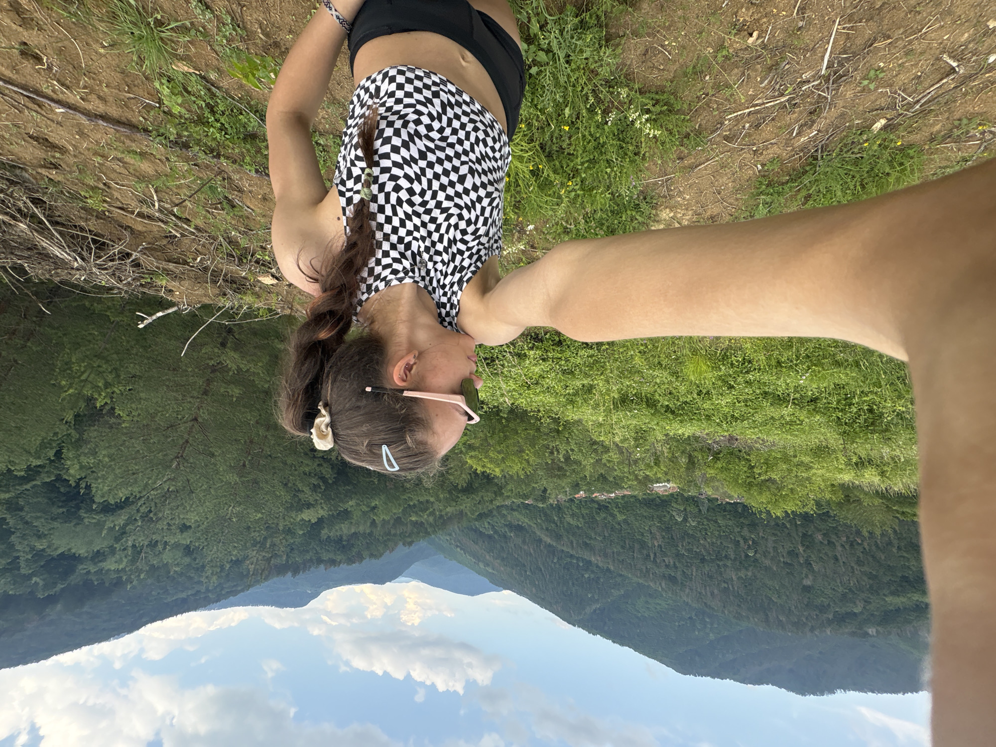 A selfie-style photograph captures a young individual, appearing to be female, on an elevated outdoor vantage point in Unknown, Unknown. Her left arm extends into the left side of the frame, holding the camera. She is positioned with her back partially to the camera, looking towards the right, observing the expansive mountainous landscape. She is dressed in a black and white checkered crop top with a wavy pattern and black shorts. Her dark brown hair, featuring lighter streaks, is styled in a long braid, secured by a cream-colored scrunchie and a light blue hair clip. She wears light pink-framed sunglasses, a stud earring in her visible left ear, and a patterned bracelet on her left wrist. The immediate foreground consists of a dirt path with scattered dry branches, sparse green grass, and small plants, sloping gently downward to the right. The midground features lush green foliage, including bushes and trees, covering a hillside. To the right, a patch of cleared land contains an accumulation of cut branches. In the distant valley on the left, a small cluster of buildings with orange-red roofs is visible. The background is dominated by multiple layers of tree-covered mountains extending into the horizon, with some areas of visible tree sparsity or defoliation on the slopes. The sky is bright blue with scattered white cumulus clouds.