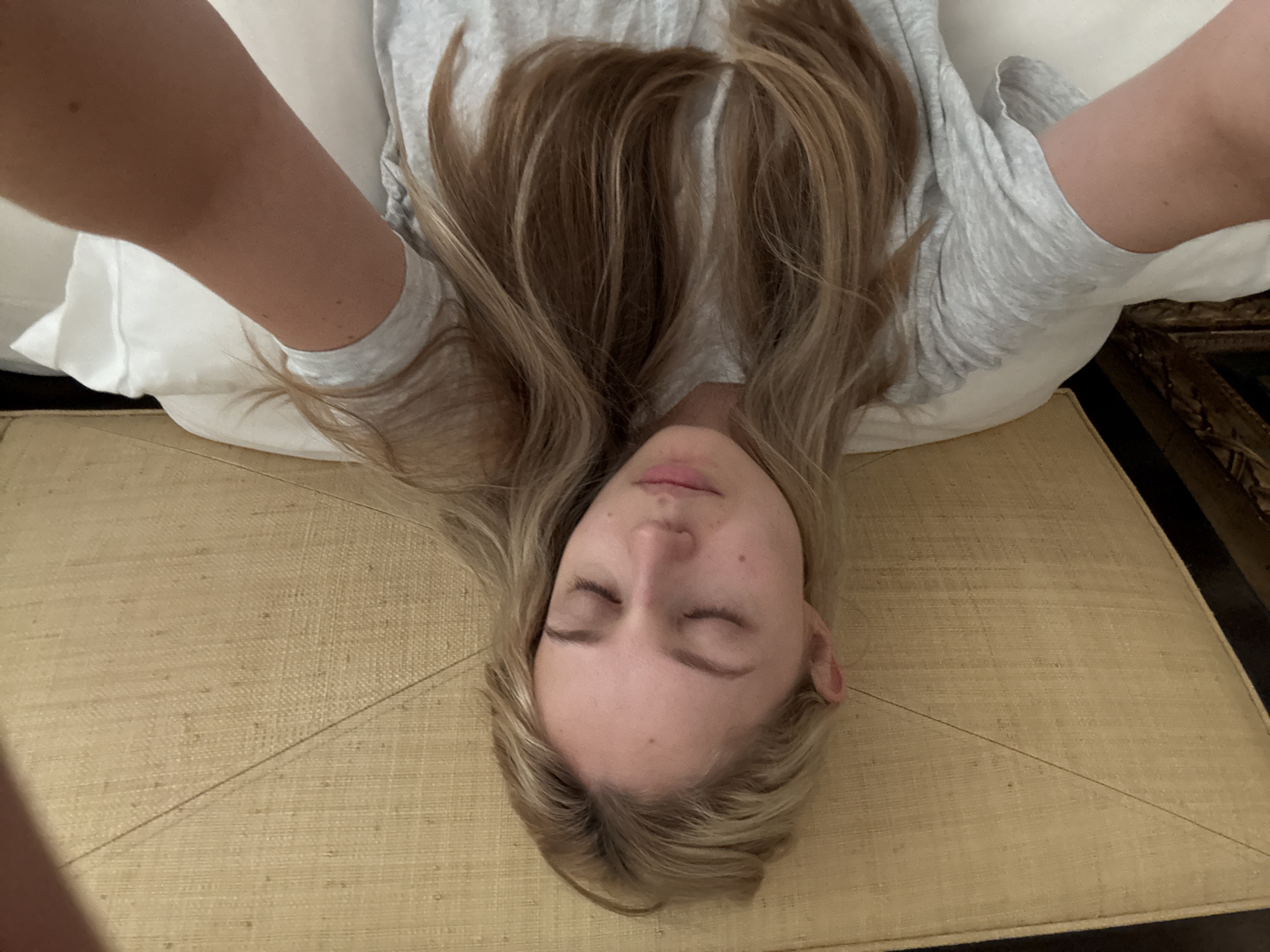 An overhead medium shot shows a woman lying in bed, her eyes closed. She has fair skin and long, blonde hair that is slightly disheveled. She is wearing a light gray t-shirt. Her head rests on a white pillow, and behind her is a headboard with a textured, woven surface. The bed appears to be in a room. There is no visible indication of the location being Ciudad de México, Mexico.