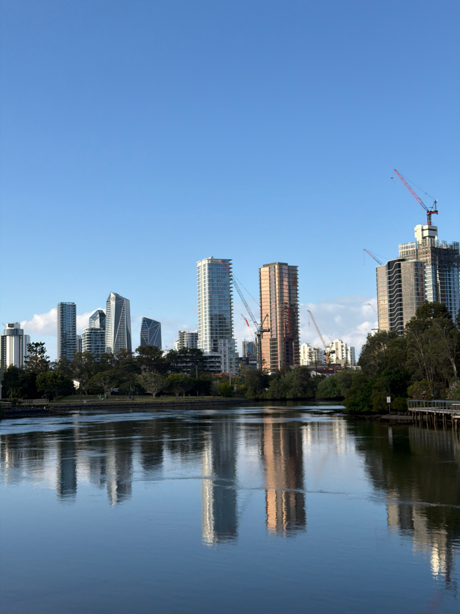 This is an image of the Broadbeach Waters skyline in Australia, captured on a clear, blue sky day. The foreground features a calm body of water, likely a canal or river, reflecting the city's architecture. Several tall, modern buildings dominate the skyline, with some appearing to be under construction, indicated by visible cranes. The buildings are a mix of glass, concrete, and a distinctive copper-colored skyscraper. Lush green trees line the banks of the water, separating the natural environment from the urban development. A wooden boardwalk or pier is visible on the right side of the image, extending over the water. The lighting suggests it might be morning or late afternoon, casting soft light and subtle reflections on the water's surface. There are no people or animals visible in the scene.