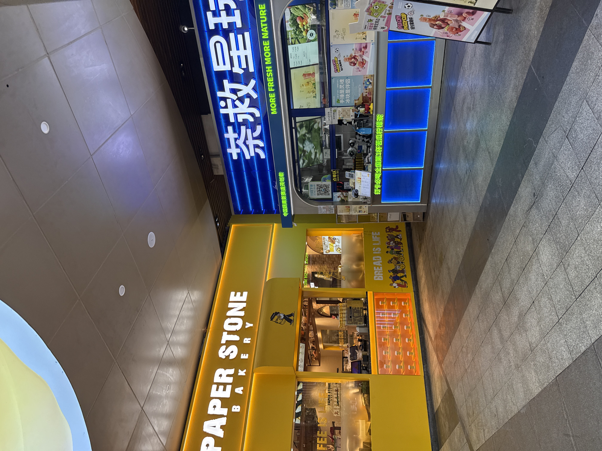 This is a medium-angle, eye-level, outdoor shot of a street scene in Haizhu District, China, featuring two commercial storefronts. The storefront on the left is yellow and displays the "PAPER STONE BAKERY" logo in white illuminated letters, with "BAKERY" in smaller letters below. The storefront on the right has a blue illuminated sign with white Chinese characters and the English text "MORE FRESH MORE NATURE". Both establishments appear to be food and beverage vendors. The pavement in front of the shops is made of grey square tiles, with darker grey sections forming pathways. Shadows from the buildings and a curved overhead structure, possibly part of a mall or covered walkway, are cast across the pavement. The lighting suggests it could be daytime with overhead artificial lighting also present. No individuals are clearly visible in the scene, though the graphics on the storefronts depict cartoon figures.