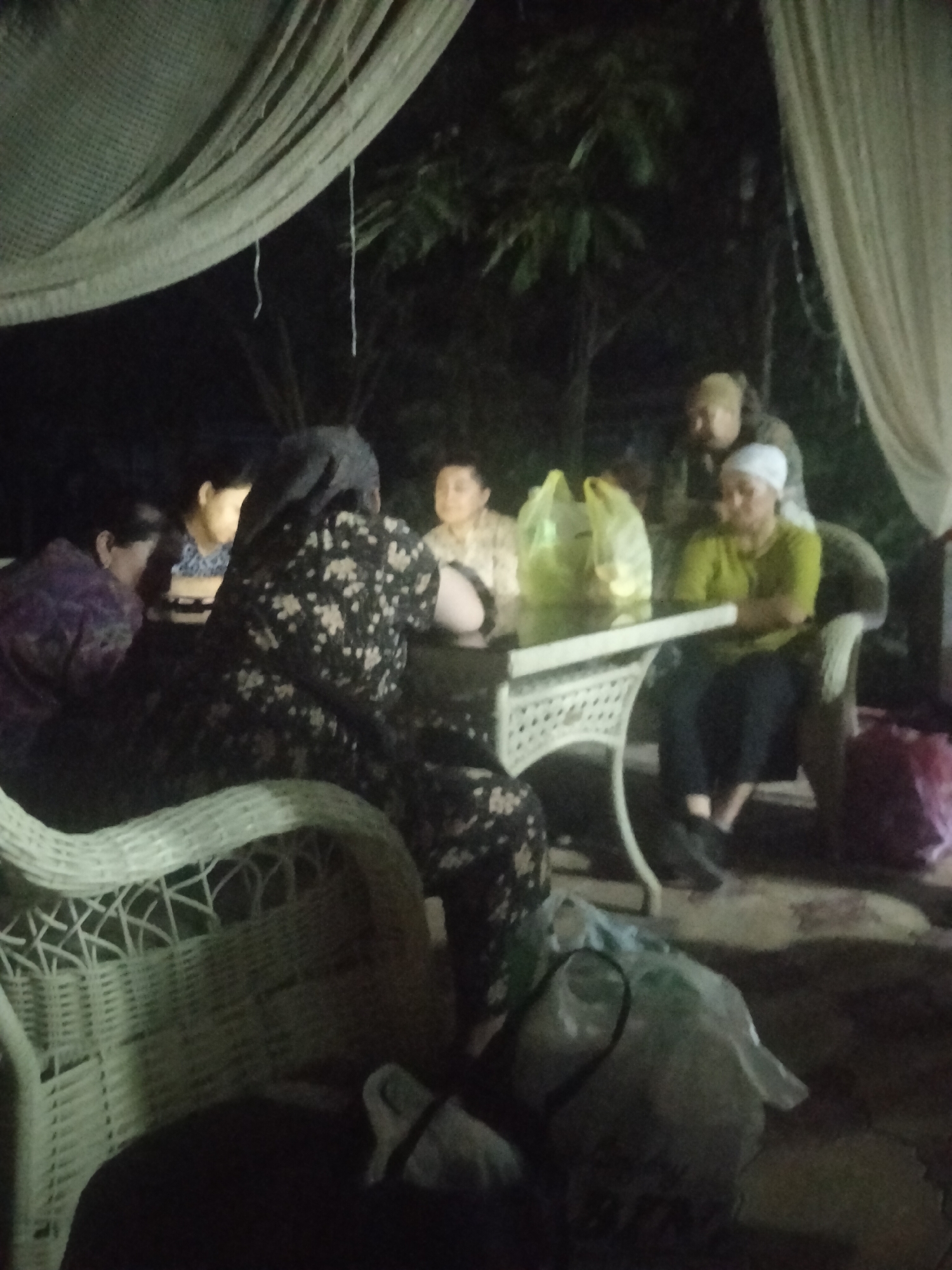 The image depicts a group of six individuals gathered around a table outdoors at night. Two women are seated on the left, one in a purple garment and the other in a black floral dress. In the center, two individuals are seated at the table, one in a light-colored shirt and another in a green shirt. Behind them, two more individuals are partially visible, one wearing a light-colored head covering and another in darker clothing. The setting appears to be a patio or garden area, with draped fabric overhead and foliage in the background. There are plastic bags on the table and a pink bag on the ground to the right. A wicker chair is prominent in the foreground on the left. The lighting is dim, suggesting an evening or nighttime scene. The image quality is low, with significant blur and low resolution, making precise identification of individuals and details challenging. The location is identified as Kagan, Uzbekistan.