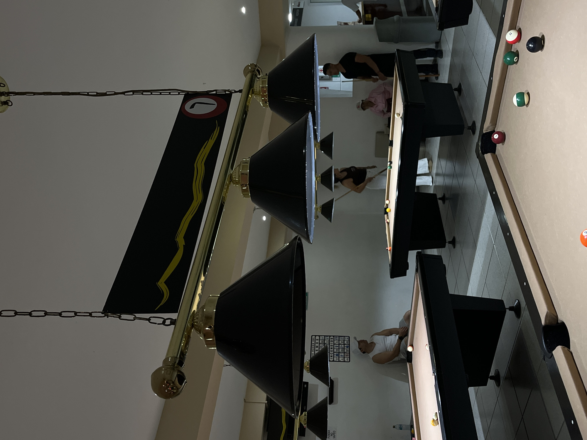 The image captures the interior of a billiards room. Several pool tables are present, with individuals engaged in playing billiards at some of them. Overhead, a light fixture with multiple lamps illuminates the tables. A black sign adorned with a yellow design and a number is suspended from the fixture. In the background, signage in Spanish is visible on a wall. The location is Unknown, Unknown.