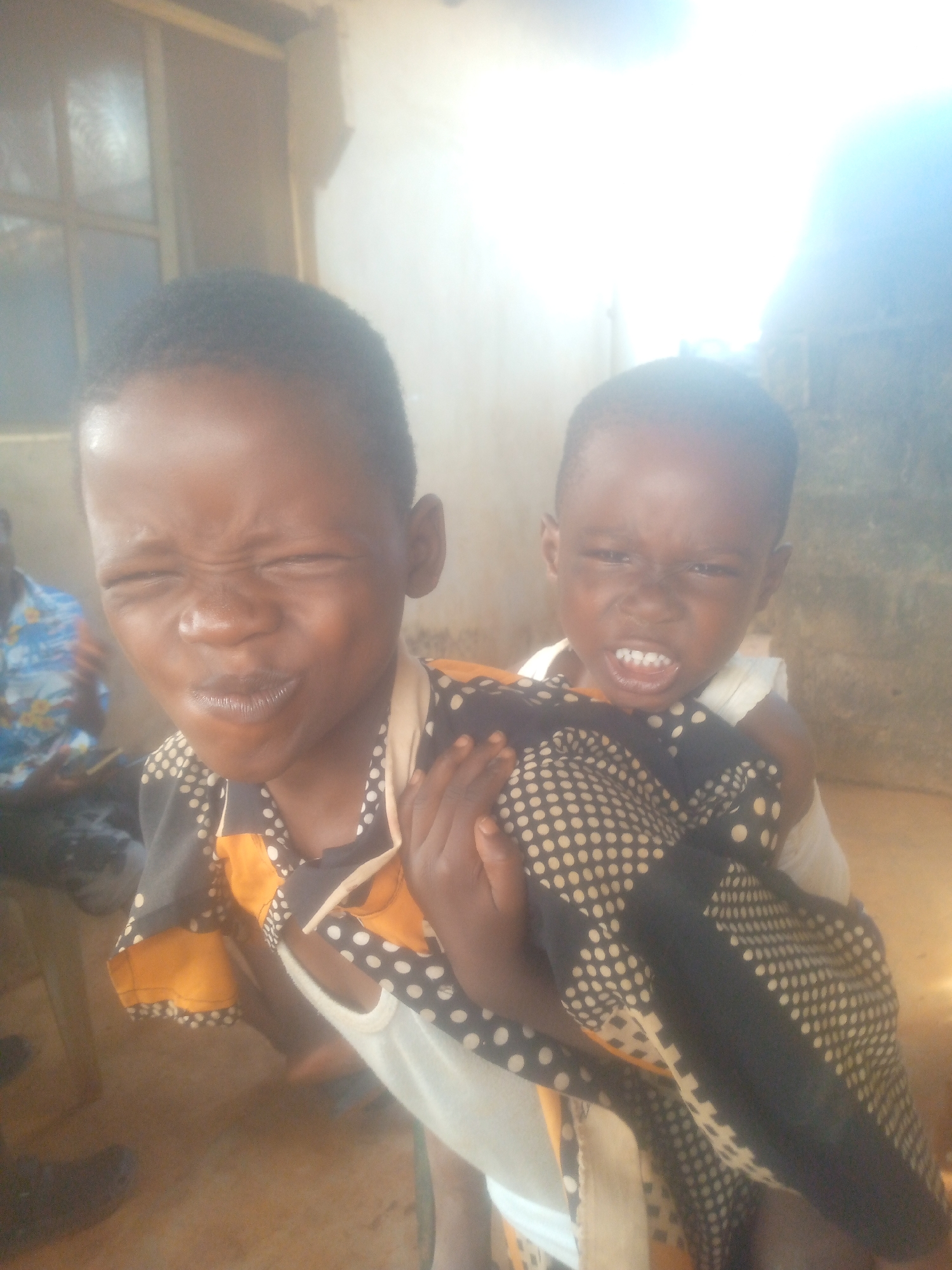 Two children are depicted indoors. The older child is carrying the younger child on their back. Both children are looking directly at the camera. The older child has their eyes squinted and is smiling slightly, while the younger child is squinting their eyes and clenching their teeth. The older child is wearing a white tank top with a patterned shirt or blanket draped over their shoulders. In the background, a person wearing a blue patterned shirt can be seen seated. The location is in the city of Unknown, Unknown.