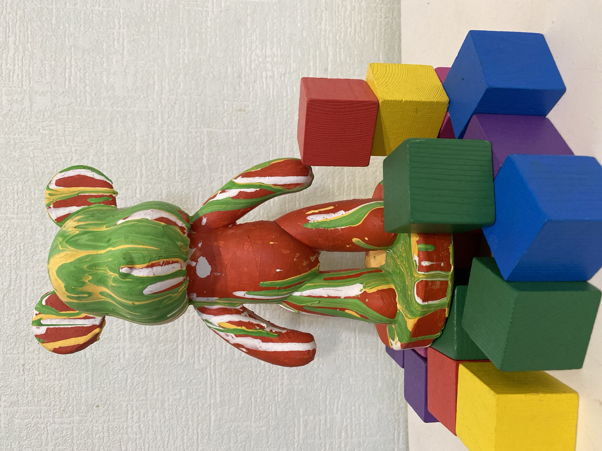 A painted toy bear with a green head and red body rests against a stack of colorful wooden blocks. The blocks are painted red, yellow, green, blue, and purple. The background is a textured white wall. The location is not evident from the image.