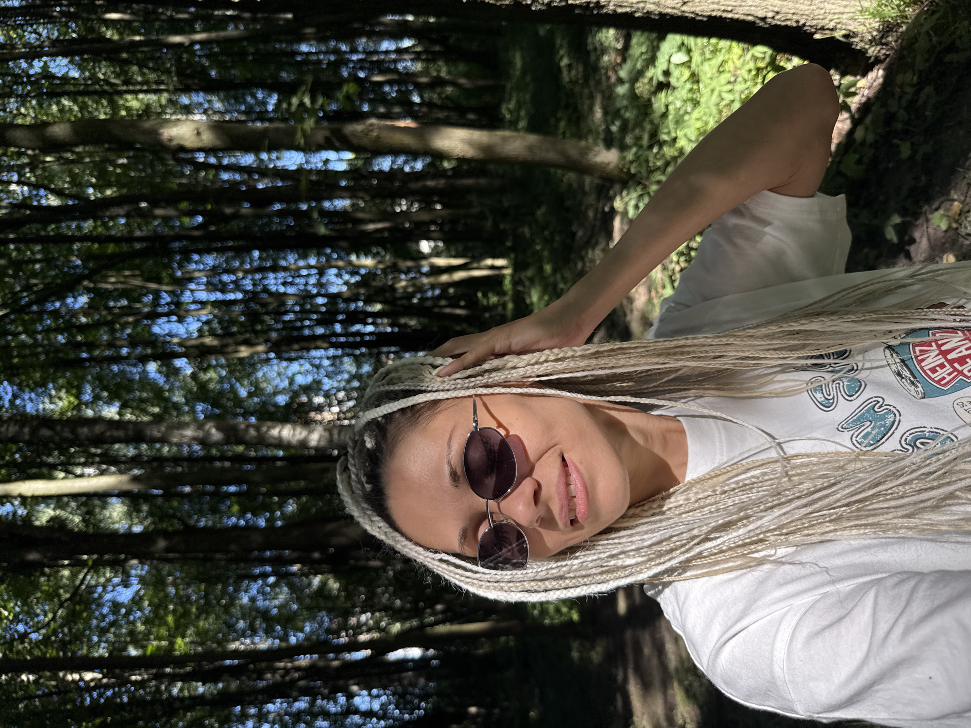 A medium shot captures a woman with blonde braided hair and sunglasses, wearing a white t-shirt, standing in a sun-dappled forest. She is looking to her left, with her right hand touching her hair. The background consists of tall trees, their trunks creating a vertical pattern, with dappled sunlight filtering through the foliage, suggesting an outdoor environment in a forest. The city of Rusanovo, Russia, is not identifiable within this scene.