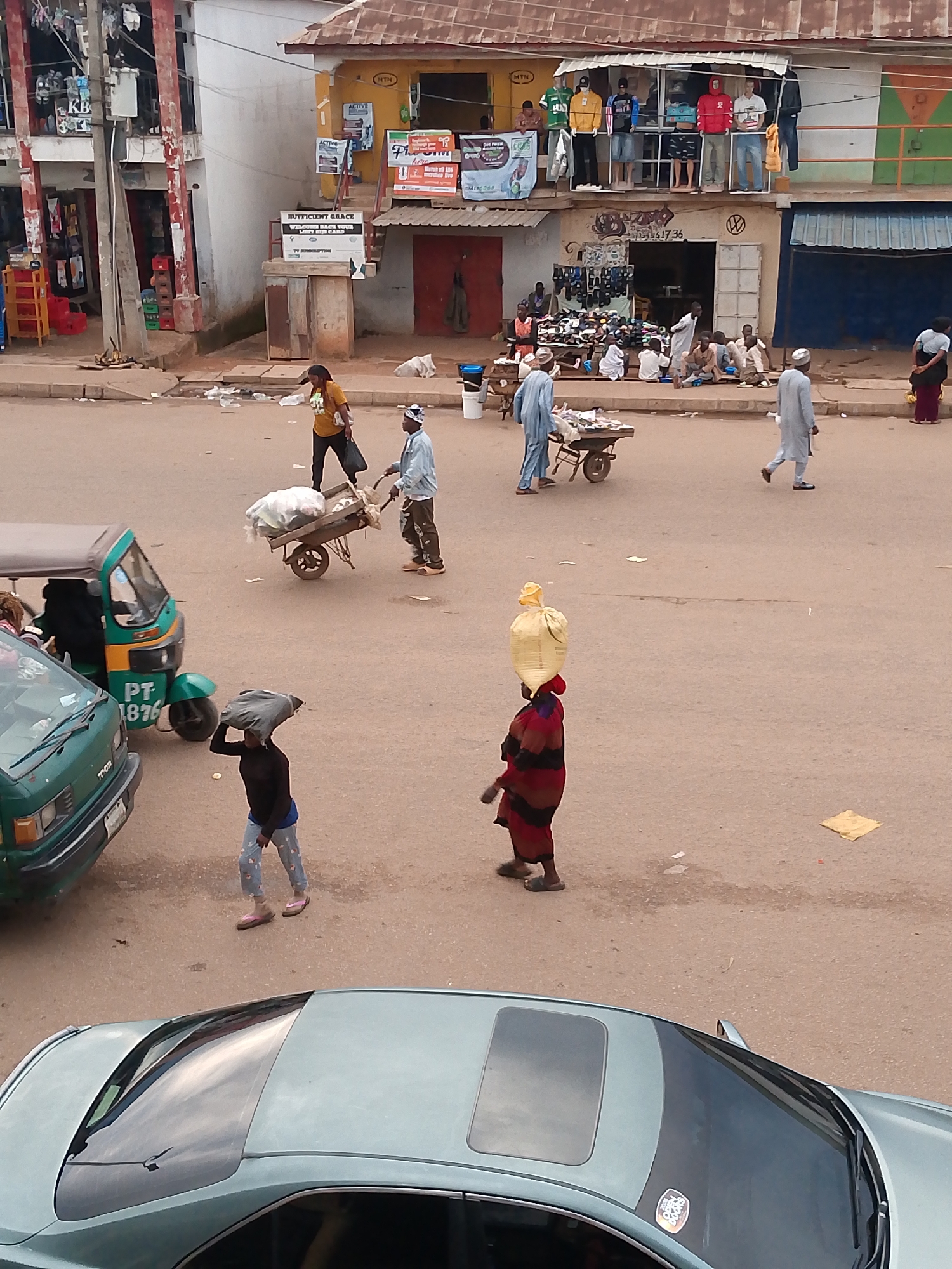 The image captures a street scene in Bukuru, Nigeria, likely a marketplace, with various individuals engaged in daily activities. In the foreground, a light blue sedan with a sunroof is parked, reflecting the sky. To its left, a green commercial tricycle (keke napep) with registration PT-1876 is visible. Further into the scene, a person in a dark top and patterned pants carries a dark gray bag over their head. A man in a light blue shirt and dark pants pushes a wooden cart filled with goods, followed by another man in a light blue robe. Two individuals in blue robes are walking away from the cart. In the background, several shops are open, displaying goods like clothing and shoes. Mannequins are visible in the upper floor windows of one shop. People are seen sitting and standing around the shops. A woman in a yellow shirt and dark pants walks away from the viewer, carrying a black bag. The ground is a paved road with scattered debris and small yellow papers. The buildings appear to be made of concrete and corrugated metal roofing.