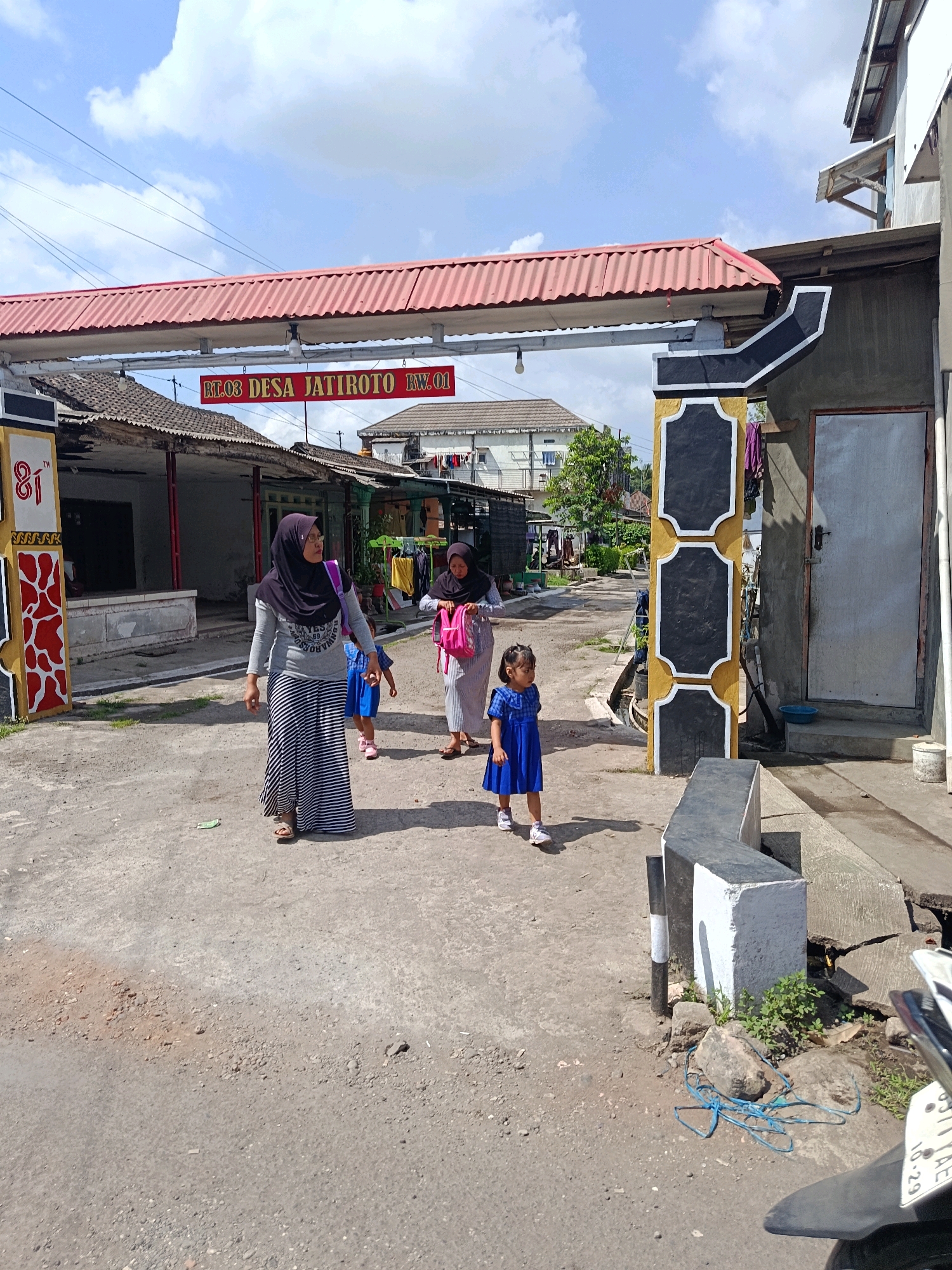 The image depicts an outdoor scene in Jatiroto, Indonesia, under a cloudy sky. In the foreground, a young girl in a blue dress is walking on a dusty road. Behind her, a woman in a gray shirt and a striped skirt, wearing a hijab, is walking. Another woman, also in a hijab and a striped dress, is walking further back. They appear to be moving away from the camera.
Overhead, a red corrugated metal awning stretches across the scene, supported by pillars. A sign under the awning reads "RT.03 DESA JATIROTO RW.01" in red letters. The background shows simple buildings with tiled roofs, some with laundry hanging outside. A motorcycle is partially visible on the right side of the image, with its license plate showing "B 711 AE" and "10.29". The ground is a mix of dirt, gravel, and cracked concrete. The overall environment suggests a residential or semi-commercial area within the village.