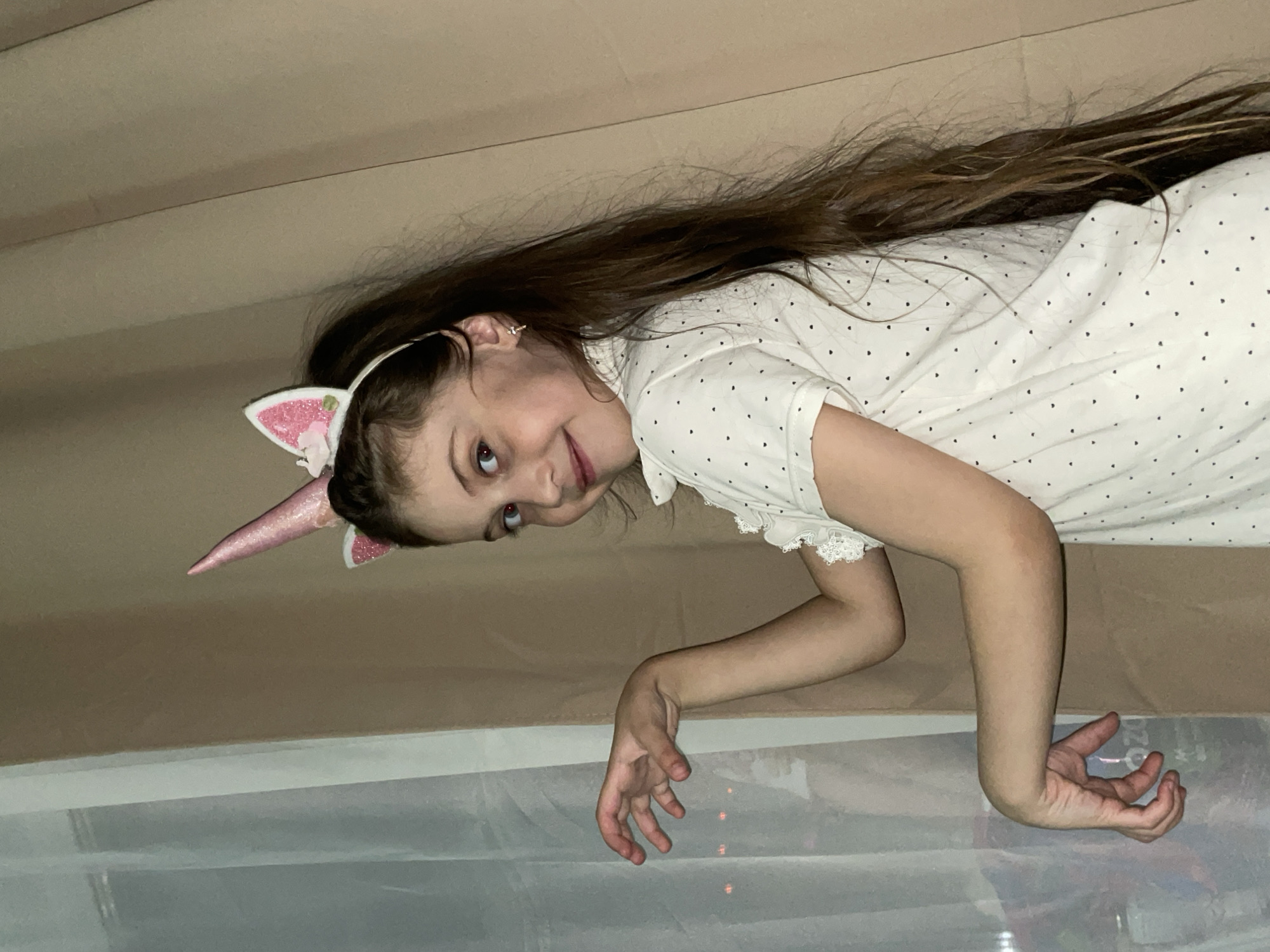 A young girl wearing a white, polka-dotted dress and a pink unicorn headband is lying on a bed. She has long brown hair. Her right arm is bent at the elbow, with her hand extending towards the lower edge of the image. The environment consists of a beige backdrop and what appears to be a clear plastic sheet. The location of the image is not provided.