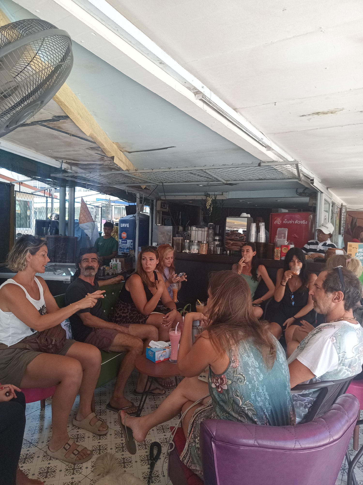 A group of people are sitting and socializing at an open-air cafe in ตำบลเกาะพะงัน, Thailand. The cafe has a tiled floor and a partially covered ceiling with exposed beams and lighting fixtures. Several people are seated in chairs around small tables, drinking beverages and engaging in conversation. A man in a green shirt stands behind a counter, possibly a waiter or barista. There's a beverage cooler with the word "WAVE" on it. Other people are seated at a counter in the background. A fan is visible in the upper left corner. A small dog is lying on the floor near the seated group. The environment appears relaxed and casual.