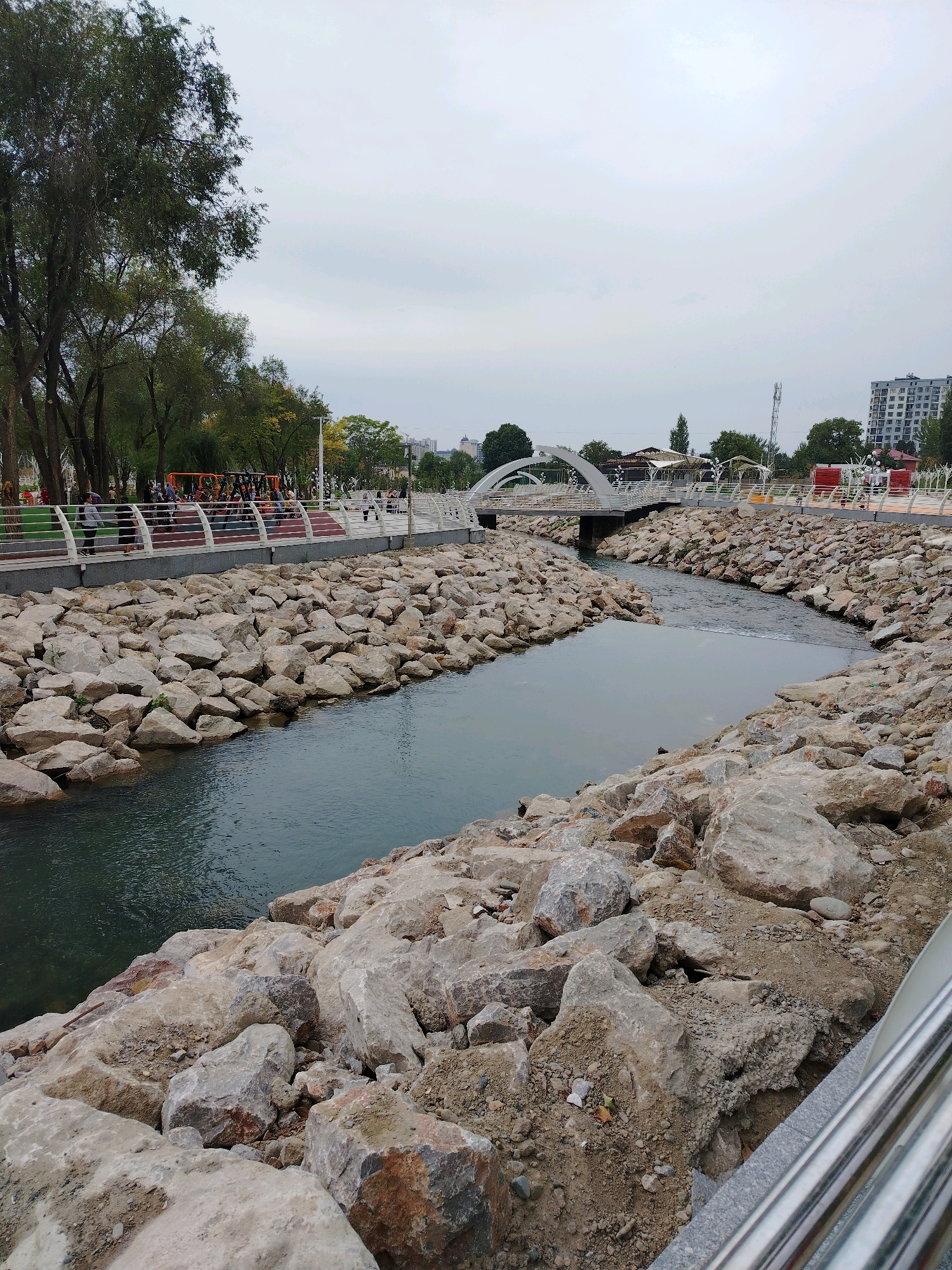 The image shows a view of a paved walkway alongside a river in Osh City, Kyrgyzstan. The walkway is lined with a metal railing and trees. There are people walking on the walkway. The river is surrounded by large rocks. A white arched bridge crosses the river in the background. Buildings can be seen in the distance. The sky is overcast.