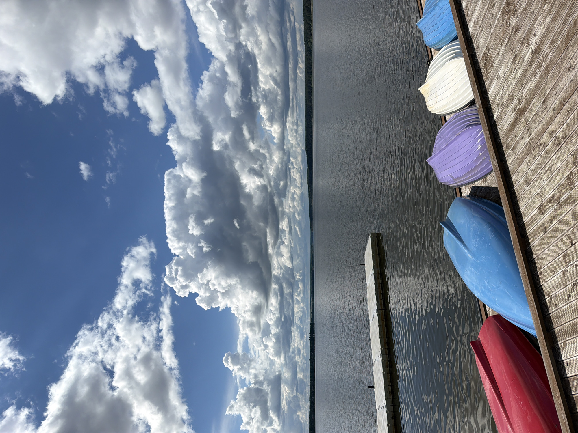 The image depicts a lakeside scene, likely a recreational area with a wooden dock extending into the water. Several small boats, perhaps rowboats or dinghies, are stacked upside down along the side of the dock. The water is calm with visible reflections. A line of trees is visible across the lake. The sky is dominated by large, puffy cumulus clouds against a blue sky. There is no indication that the location is in Votkinsk, Russia. No people or actions are visible.
