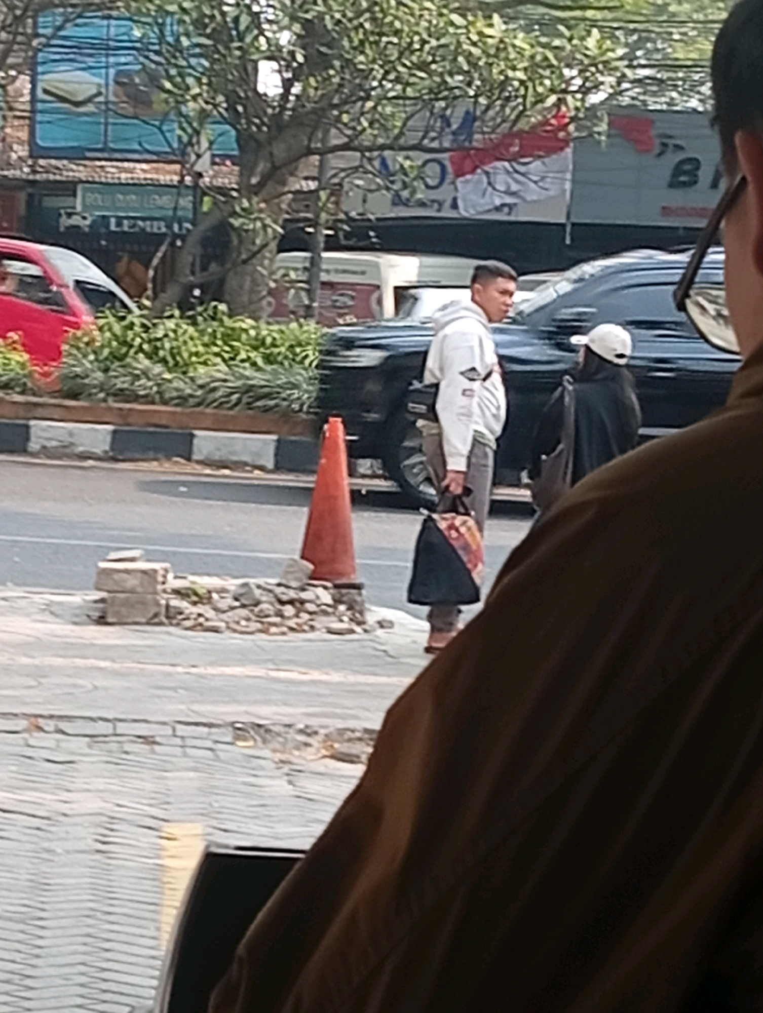 A medium shot captures a street scene in Bandung City, Indonesia, on a sunny day. In the foreground, the dark brown fabric of a jacket fills the right side of the frame, obscuring much of the viewer's perspective. A paved sidewalk with a brick pattern runs horizontally across the bottom of the image. To the left of the sidewalk, a section of asphalt road is visible, marked with a white line and a reflective yellow strip.
On the sidewalk, a bright orange traffic cone stands near a small pile of bricks and rubble. Further to the left, a red car is partially visible, parked or moving. Behind the cone and car, a dark-colored SUV is the dominant vehicle, its glossy surface reflecting the surroundings.
Two individuals are in the mid-ground, positioned on the sidewalk near the SUV. A man, wearing a white hoodie and grey pants, stands facing left, carrying a patterned bag in his right hand. He appears to be looking away from the camera. Beside him, a person wearing a dark hooded jacket and a white baseball cap is seen from the back, also facing left and holding a bag.
In the background, a large tree with dense green foliage partially obscures a building. The building features a large advertisement or mural with red and white elements, possibly a flag or a banner, and text that is difficult to decipher. A street curb with black and white markings separates the road from a strip of green bushes. The overall scene suggests an urban environment, likely a busy street with pedestrian and vehicle traffic.