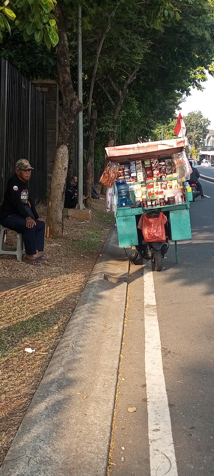 A man wearing a camouflage hat, black long-sleeved shirt, and dark pants sits on a grey plastic stool by a metal fence. He is facing left, with his hands in his lap. Behind him, a small green cart, mounted on a motorcycle, is filled with various packaged goods and water bottles. A red plastic bag hangs from the cart. To the right of the cart, a motorcycle is being ridden away from the viewer. In the background, trees line the street, and a white and red flag is visible on top of the cart. The scene appears to be on the side of a road in Lambangjaya, Indonesia, with a sidewalk separating the street from a grassy area and the fence.