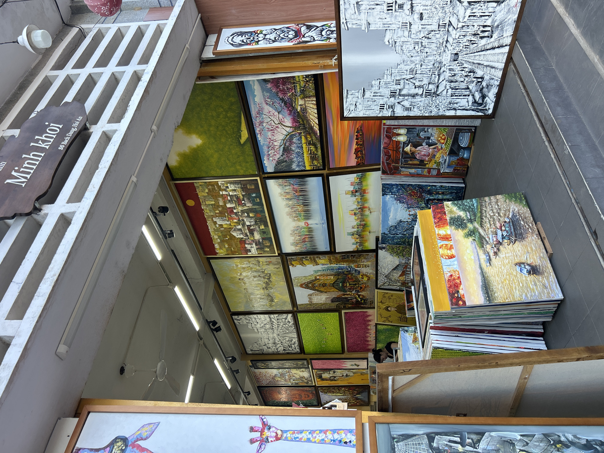 This image captures the open entrance of an art gallery or shop identified by a wooden sign overhead reading "Minh khoi" and "3G Bach Dang, Hoi An." The shop is located in Hội An Ward, Vietnam.
Inside, a single individual with dark hair, wearing a dark top, is seated behind a table or counter, visible in the mid-left portion of the frame. The shop walls are extensively covered with numerous paintings of various sizes and styles, arranged in a dense, grid-like display. Visible artwork includes diverse cityscapes (some monochrome, some colorful), abstract landscapes, a painting featuring cherry blossom trees, scenes with boats on water, and at least one depiction of a street vendor. A tall, slender painting with stacked abstract figures is displayed vertically on the far right wall. A colorful giraffe portrait is partially visible on a canvas in the foreground on the left.
The shop interior features a white ceiling with multiple fluorescent tube lights and track lighting fixtures. A white four-bladed ceiling fan is mounted centrally. The floor is covered with dark gray tiles, leading up a slight step from the exterior. In the foreground and extending into the shop, stacks of paintings, both framed and unframed, are densely arranged on the floor, showcasing a wide array of subjects.
Above the entrance, a concrete structure with rectangular cutouts forms a decorative facade. To the right of the entrance, an ornate red lantern and a white cylindrical light fixture are suspended.