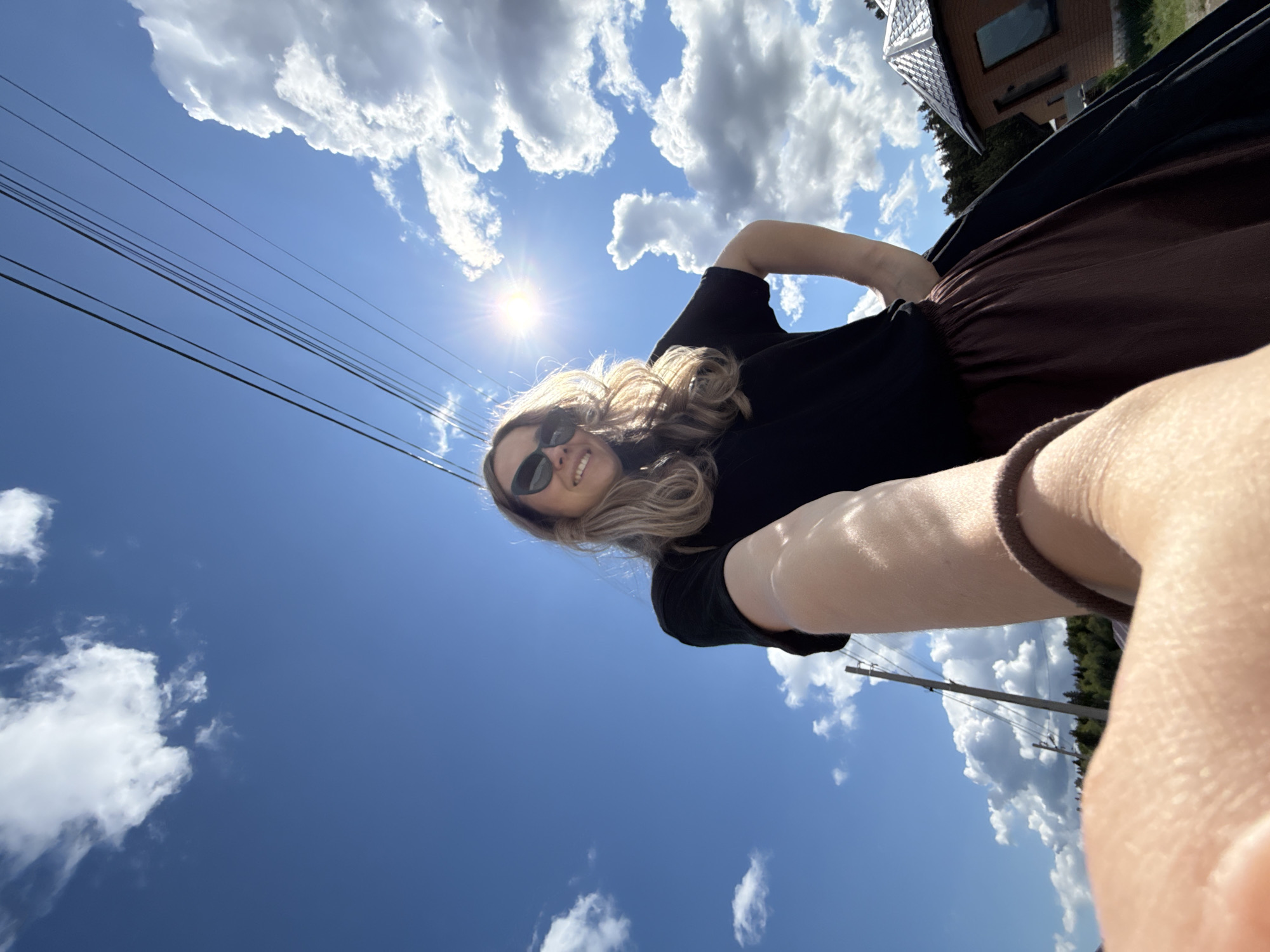 A person is taking a selfie against a backdrop of a bright blue sky with fluffy white clouds. The sun is visible in the sky, and there are power lines running across the upper-left portion of the frame.
The person has blond hair and is wearing sunglasses. They are dressed in a black top and brown pants or a skirt.
The setting appears to be outdoors, with glimpses of buildings and trees in the background. Without additional information, precise determination of the location in Chaikovsky, Russia, is not feasible.