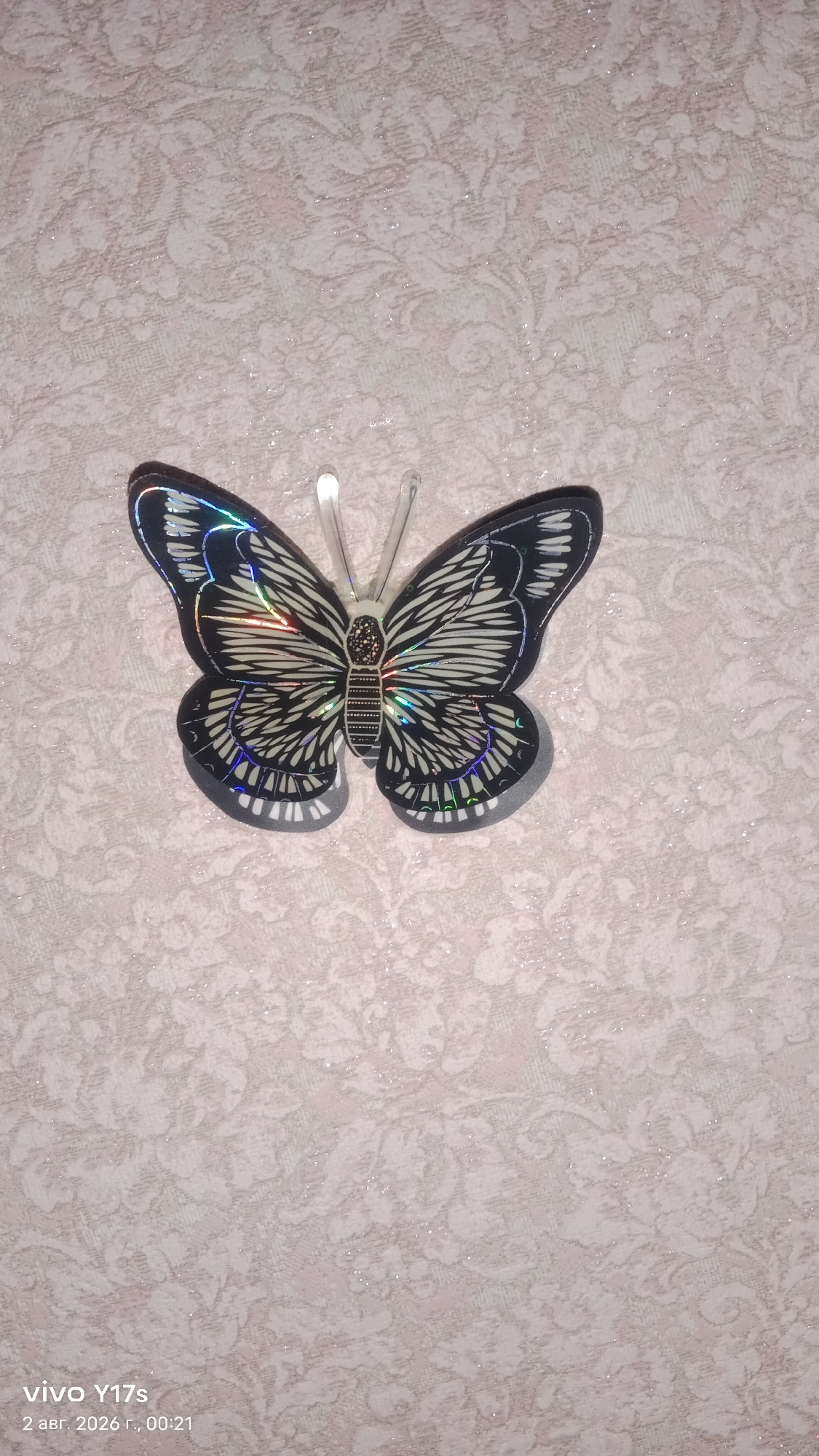 A holographic butterfly sticker is affixed to a textured, light-colored wall with a floral pattern. The sticker is centrally located in the frame, facing left. The background wall exhibits a subtle shimmering effect. The image is a photograph captured with a vivo Y17s phone on August 2, 2026, at 00:21. The location is identified as Rudny, Kazakhstan.
