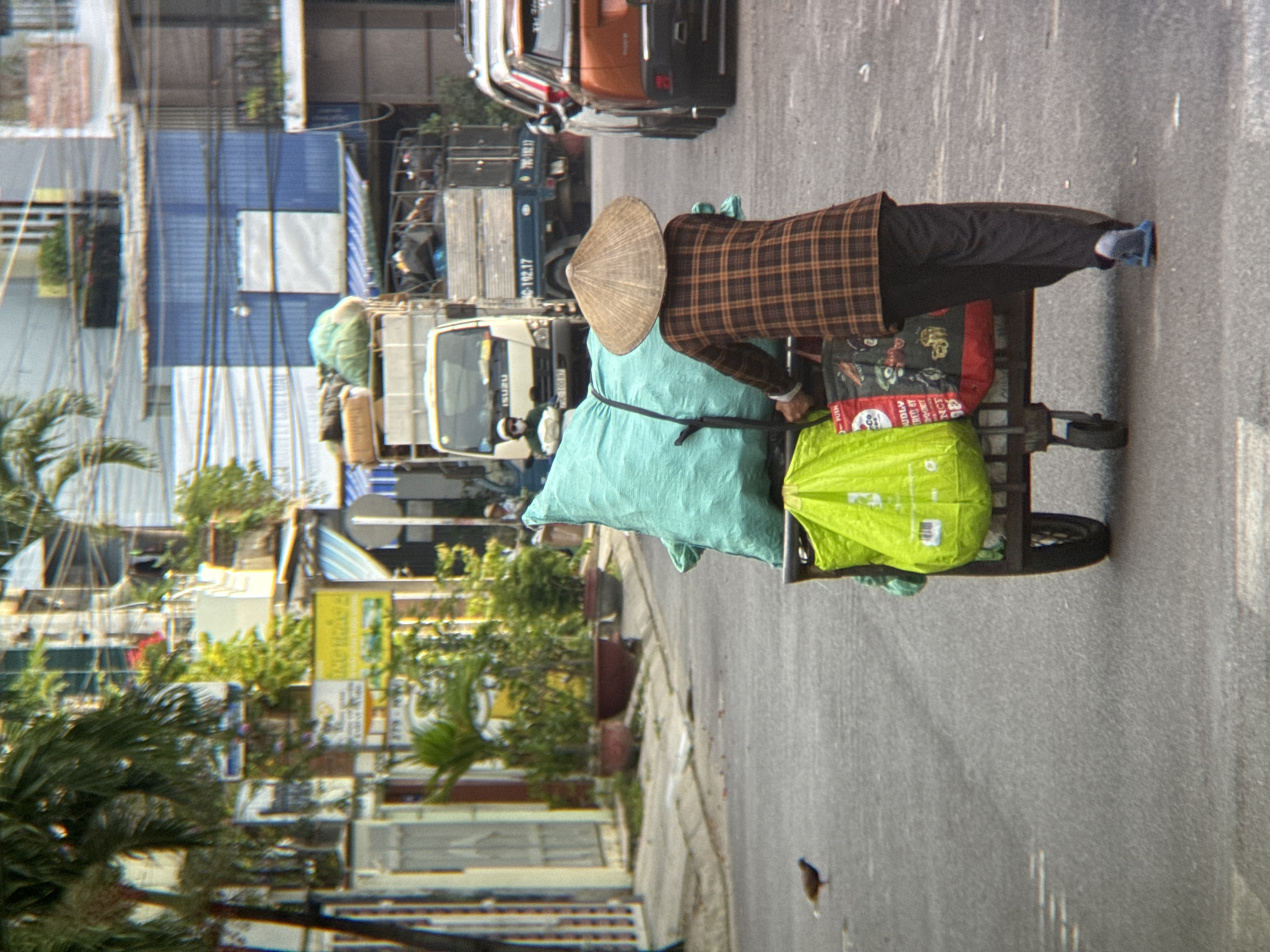 The image depicts a street scene in Nha Trang, Vietnam, featuring a person pushing a cart laden with goods. The individual, from behind, wears a traditional conical hat and a plaid long-sleeved shirt, along with dark pants and blue sandals. The cart is covered with large green and blue tarps, concealing its contents, and it also carries bright green shopping bags. In the background, various buildings line the street, some with blue and white facades, and trees are visible. Traffic includes a white truck with "ISUZU" branding, a blue truck, and a section of an orange car on the right side. A small bird is present on the asphalt in the lower left quadrant of the image. The overall atmosphere suggests a commercial or residential street in an urban environment.