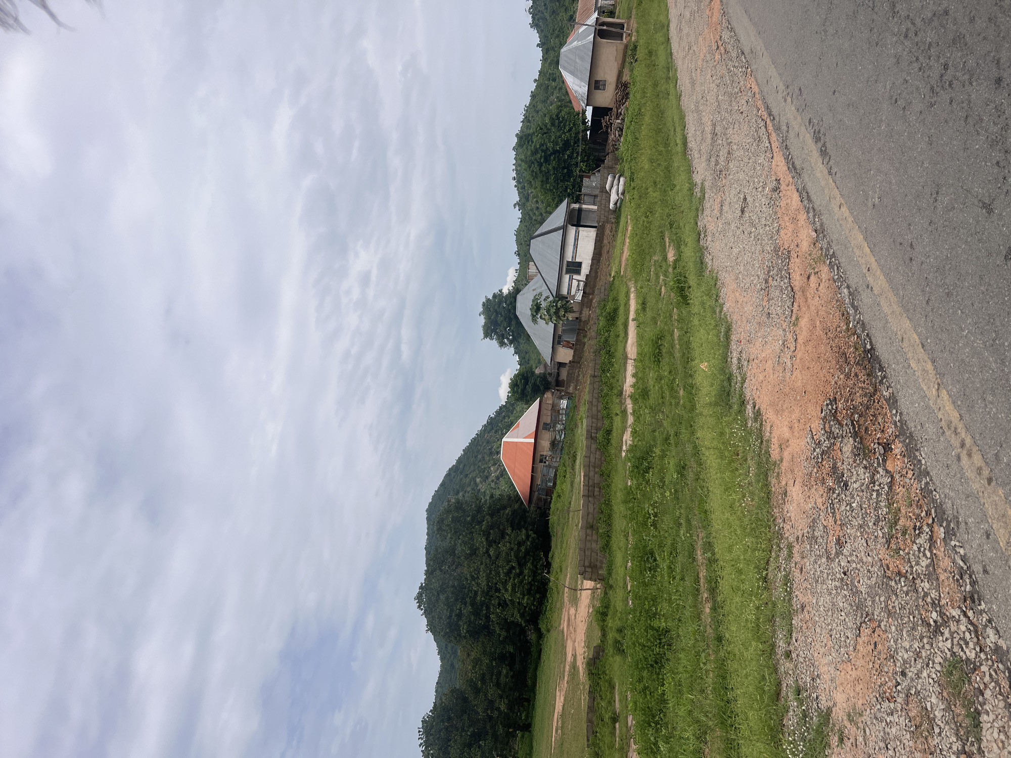 The image depicts a residential scene in Minda, Nigeria, under a cloudy sky, suggesting it's likely daytime. The foreground features a paved road with a gravel shoulder, bordered by lush green grass. A dirt path runs alongside the grass, leading towards several houses.
The houses are of varying architectural styles, with some appearing to be under construction. One house has a distinctive orange, pyramid-shaped roof. Another is a more modern-looking building with a silver, sloped roof and white walls. Behind the houses, a dense, green forested hill rises steeply, creating a striking natural backdrop.
The overall impression is one of a developing rural or semi-urban area. There are no people visible in the image. The weather appears to be overcast, but not actively raining. The scene is tranquil, with no indication of any specific activity or event taking place. The presence of the road and the houses indicates a settled community.
