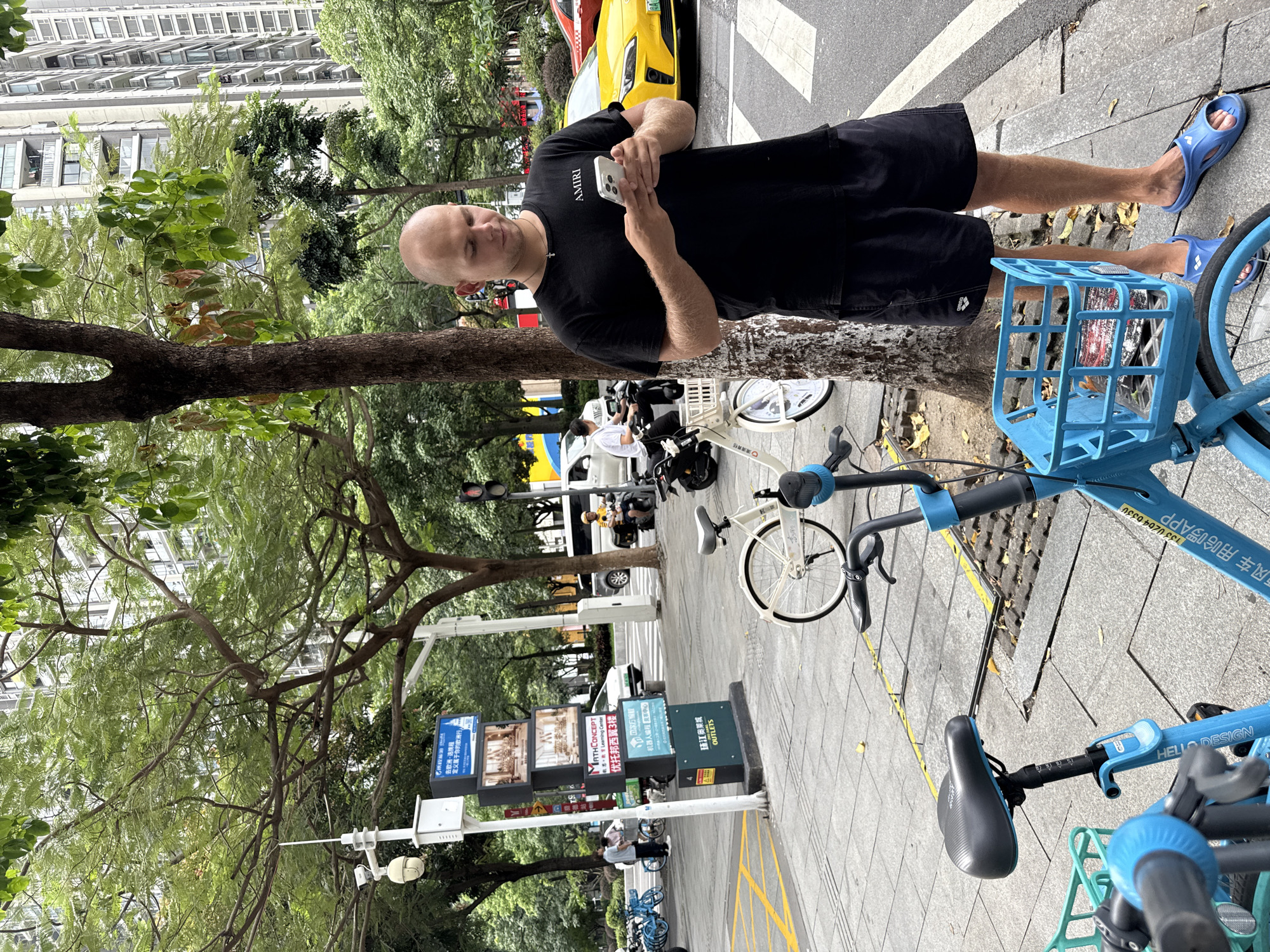 In Tianhe District, China, a bald Caucasian male wearing a black t-shirt with the "AMIRI" logo and black shorts stands on a sidewalk next to a tree, looking at his white smartphone. He is wearing blue slip-on sandals. In the foreground, a blue shared bicycle with a basket is visible, and another white shared bicycle is positioned behind it. Across the street, a yellow car and a white van are stopped at a traffic light, with a person on a scooter in front of the van. A digital billboard with multiple screens displays advertisements and information. The environment includes trees, buildings, and a paved sidewalk with yellow markings.