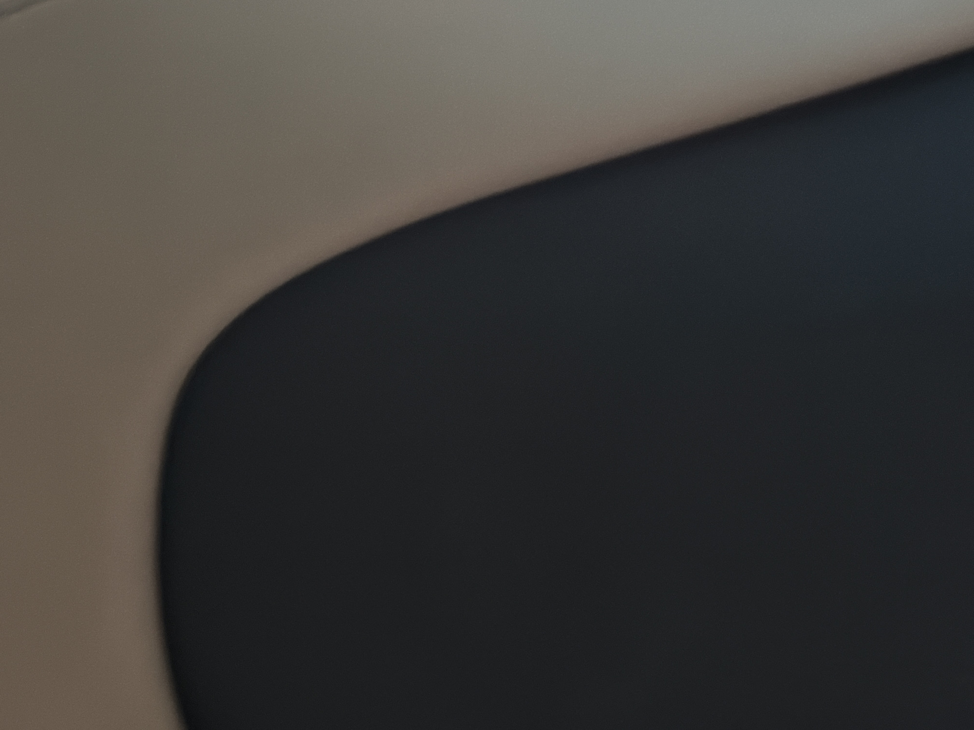 The image shows a close-up view of what appears to be the interior of a vehicle, specifically a car seat. The seat is divided into two distinct sections: a dark grey or black lower portion and a lighter beige or tan upper portion. The transition between the two colors is smooth and curved, suggesting a contoured design. The lighting in the image is soft and diffused, creating subtle shadows and highlights on the textured surface of the seat. The background is out of focus, further emphasizing the texture and color of the car seat. There are no people or other objects visible in the frame. The location is not discernible from the image itself, and therefore, it is not possible to determine if it is in Prysivtsi, Ukraine, or any other specific city.