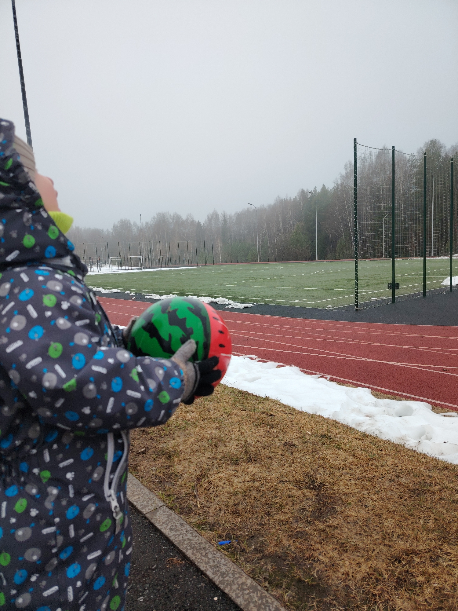A young child in a patterned winter jumpsuit and mittens is outdoors, holding a ball colored like a watermelon, looking up. The child is positioned near a running track, with patches of snow nearby. Beyond the track is a green sports field with soccer goals and a tall fence along the edge. In the background are bare trees and a light-colored, overcast sky. The location is likely in the city of Марковский, Russia.