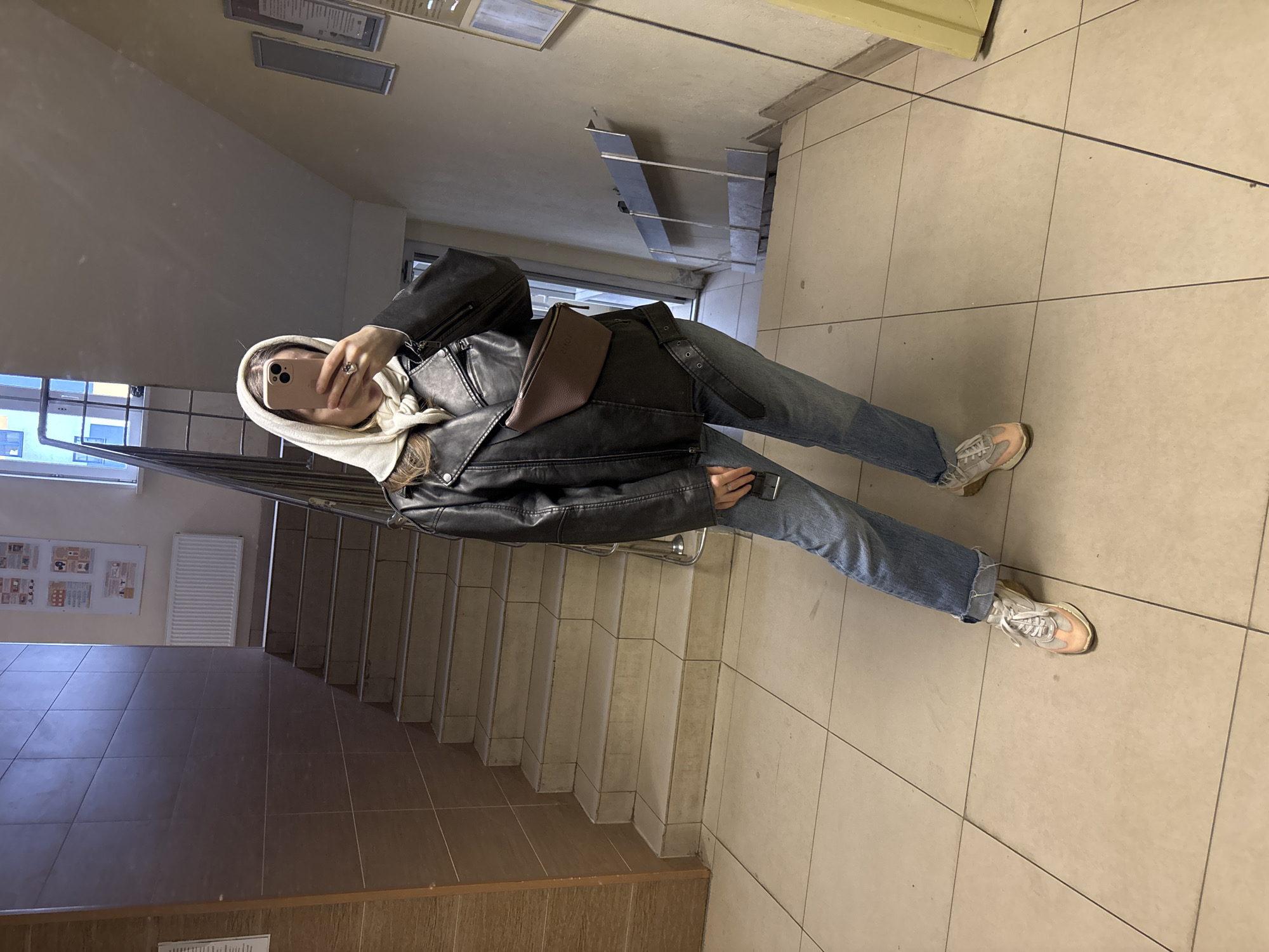 The image shows a person taking a selfie in what appears to be the stairwell of a building. The person is wearing a white hoodie, a black leather jacket with a brown fanny pack, blue jeans, and light-colored sneakers. The stairwell has light-colored tiled floors, a metal handrail, and concrete steps. A bulletin board with various papers and a white radiator can be seen on the wall near the stairs. The location is given as Kudrovo, Russia.