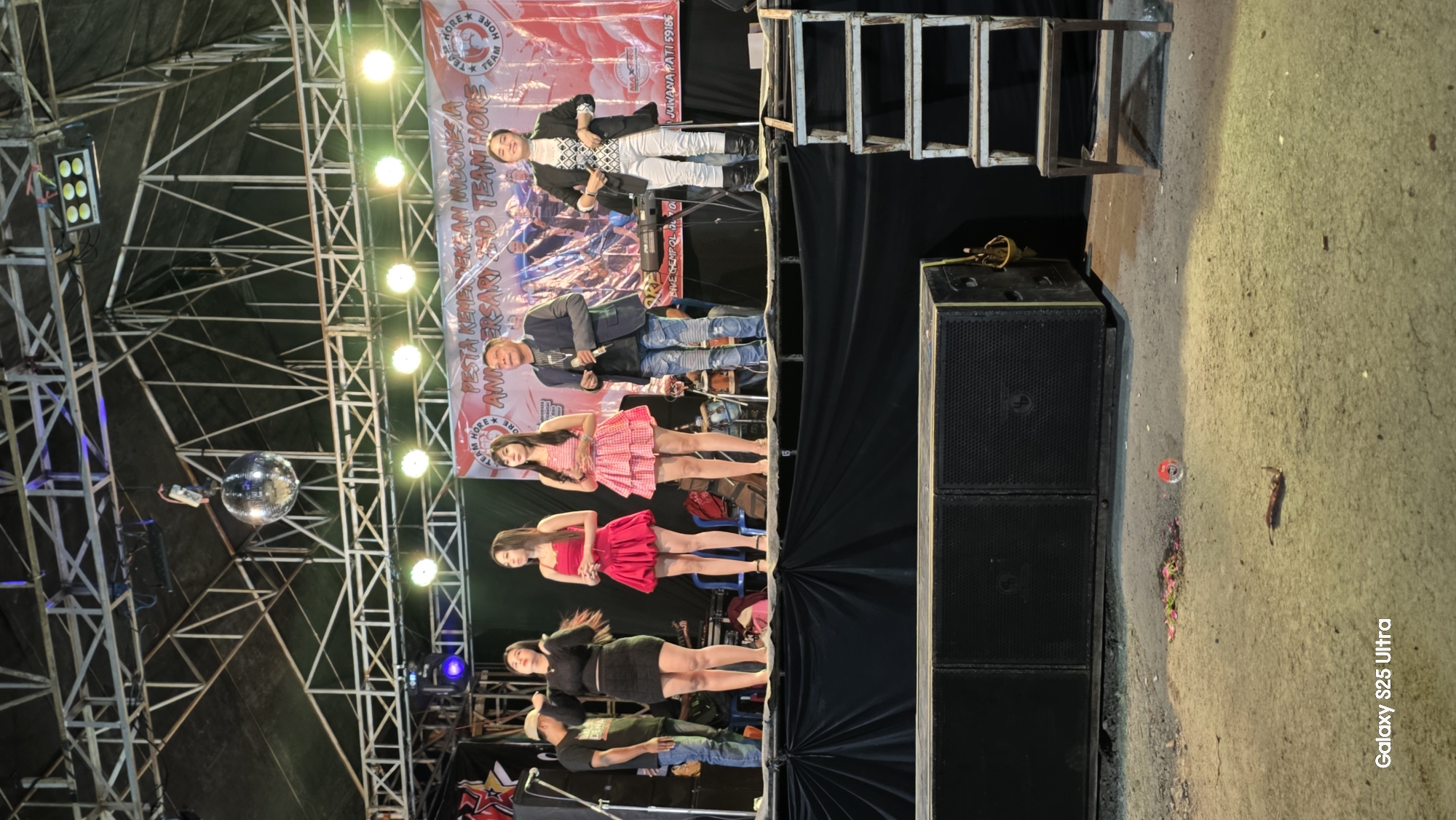 A group of five individuals, four women and two men, are performing on an outdoor stage in Juwana, Indonesia. The women are dressed in brightly colored dresses, while the men are in casual attire. They are standing on a raised platform in front of a large banner that reads "Pesta Kemerdekaan Indonesia Anniversary." A disco ball hangs from the ceiling above the stage, and lights illuminate the performers. To the right of the stage, a metal staircase leads down to the ground. In the foreground, a large speaker system is visible. The ground is rough and appears to be dirt or gravel, with some scattered debris. The overall atmosphere suggests a festive outdoor event.