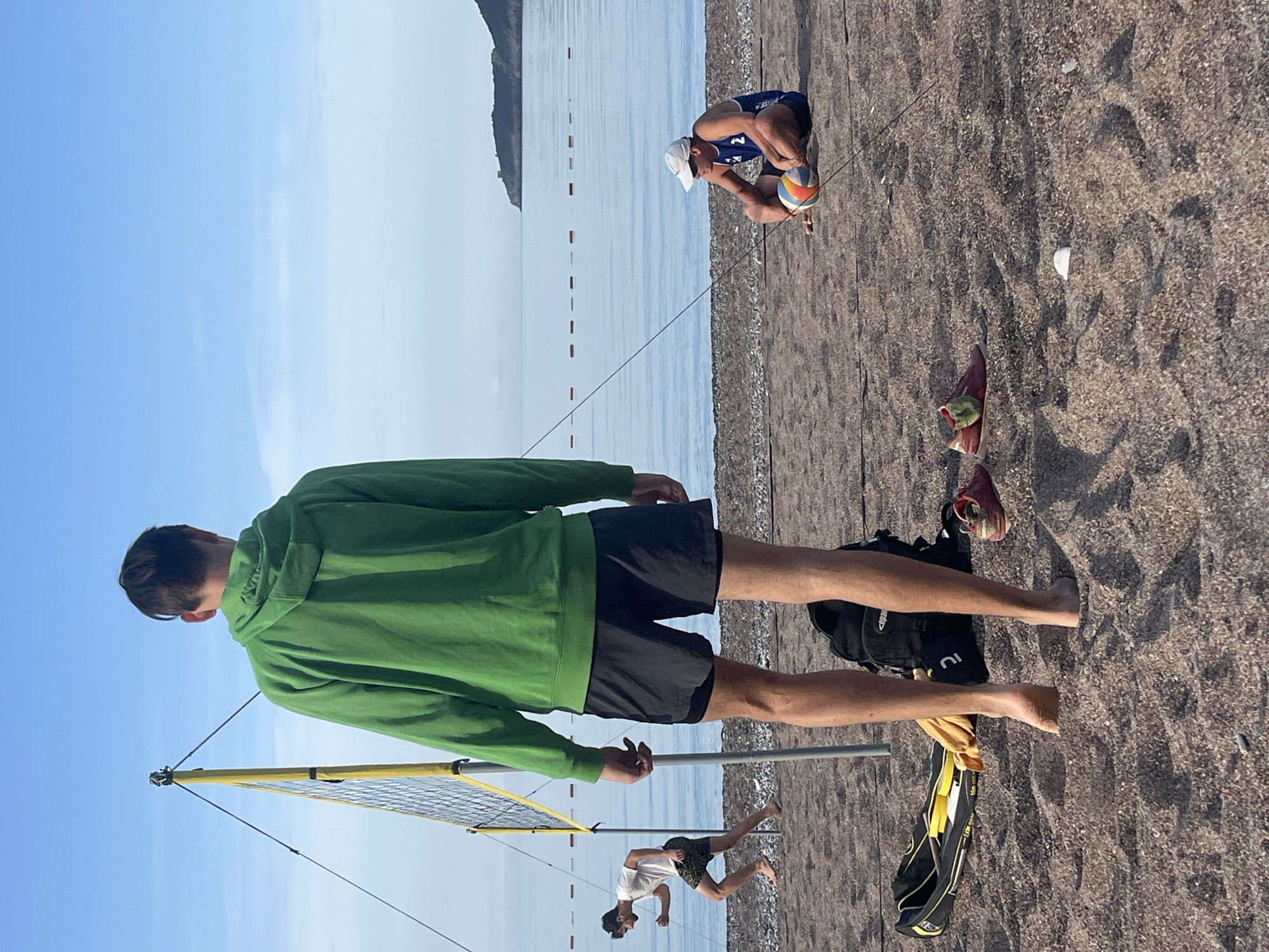 The image shows a beach volleyball scene in Boreti, Montenegro. In the foreground, a person wearing a green hoodie and black shorts stands facing the volleyball net. Behind them, on the beach, are a black backpack, a yellow bag, and a pair of red shoes. Further back, a person in a white shirt and patterned shorts is running. In the background, a person wearing a white cap and blue shorts is holding a volleyball. The sea is visible with a mountain range in the distance. Floating rectangular objects can be seen in the sea, appearing to be buoys. The sky is partially cloudy.