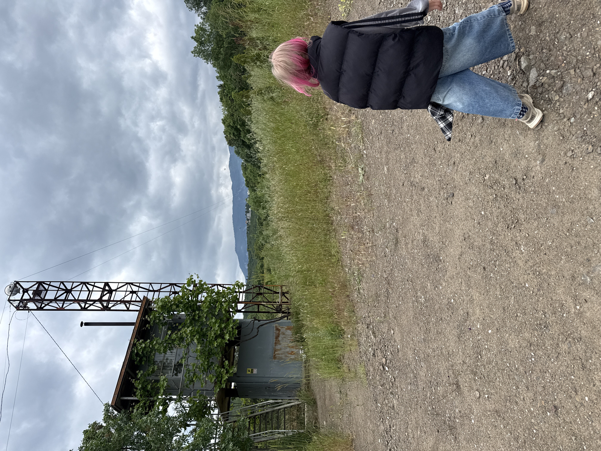 A full shot depicts an individual lying on a dirt path in an ambiguous location. The person is wearing blue jeans, a black puffer jacket, and light-colored sneakers, and has pink-tipped hair. Their posture is casual, with legs crossed and torso slightly turned toward the camera.
In the background, to the left, is a weathered, industrial structure consisting of a metal building and a supporting tower-like frame. Overgrowth of greenery is visible around the structure. The foreground consists of a dirt path, and beyond this is a grassy area. In the far distance, a mountain range is visible under a cloudy sky. The city and specific location are unknown.