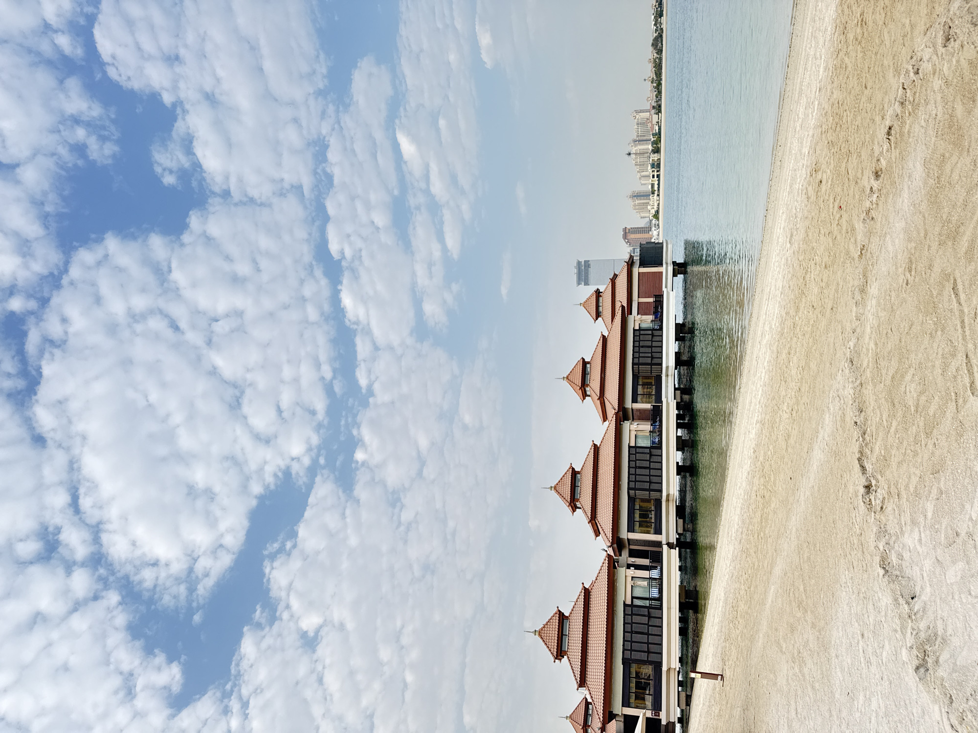This is a horizontal, eye-level, medium shot of overwater bungalows on a sunny day. The bungalows are modern with orange-tiled, pagoda-style roofs and dark-paneled windows, built on stilts over calm, blue water. A sandy beach with footprints is in the foreground, leading to the water. In the background, across the water, is a city skyline with tall buildings and palm trees, characteristic of Dubai. The sky is a bright blue with scattered white clouds.