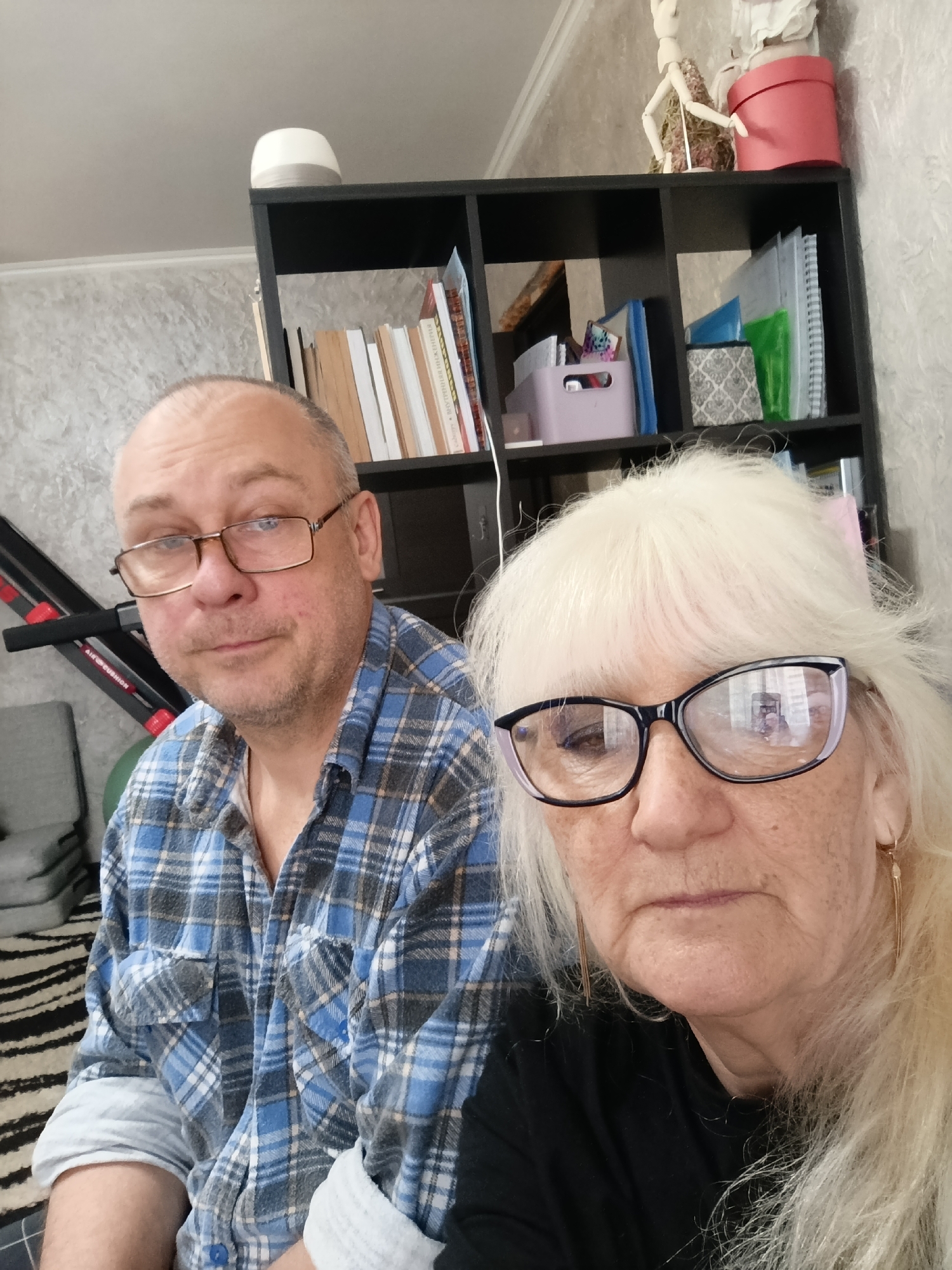 A selfie depicts two individuals indoors. A middle-aged man is on the left, wearing glasses and a blue plaid shirt. A woman with platinum blonde hair and glasses is on the right, wearing a black top. In the background is a dark shelving unit with books, notebooks, and decorative items, including a mannequin. An exercise machine and a gray cushion are visible on the left side. The location is likely a home interior. There is no information in the image to suggest the location is in Krasnodar, Russia.