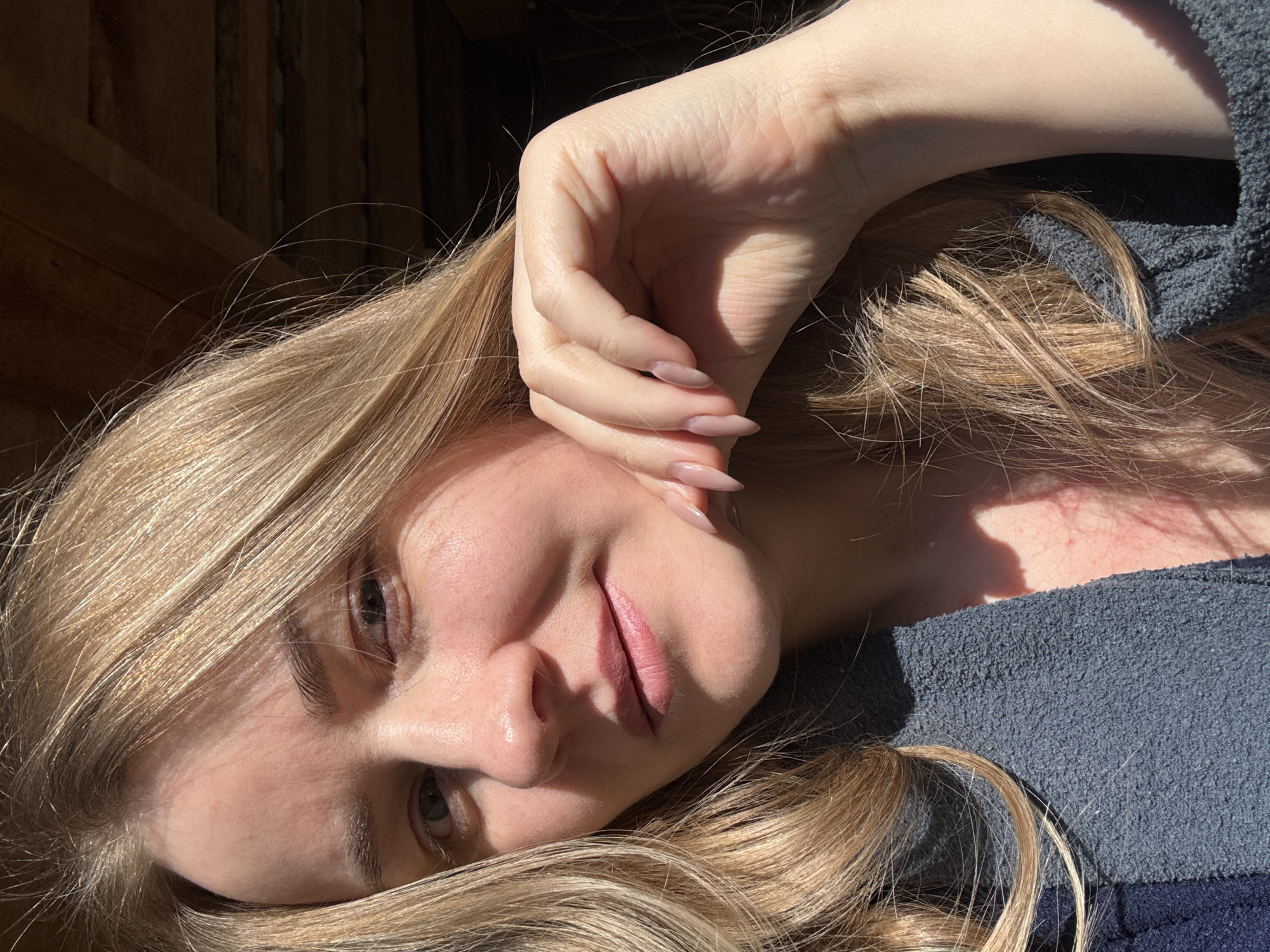 A woman with long blonde hair is lying down, looking at the camera. Her right hand is raised near her face. She is wearing a dark gray, textured garment. The background appears to be a wooden wall. The image has strong lighting, with areas in direct sunlight and others in shadow. It's impossible to ascertain if the photo was taken in the city of Chaikovsky, Russia, or any other specific location.