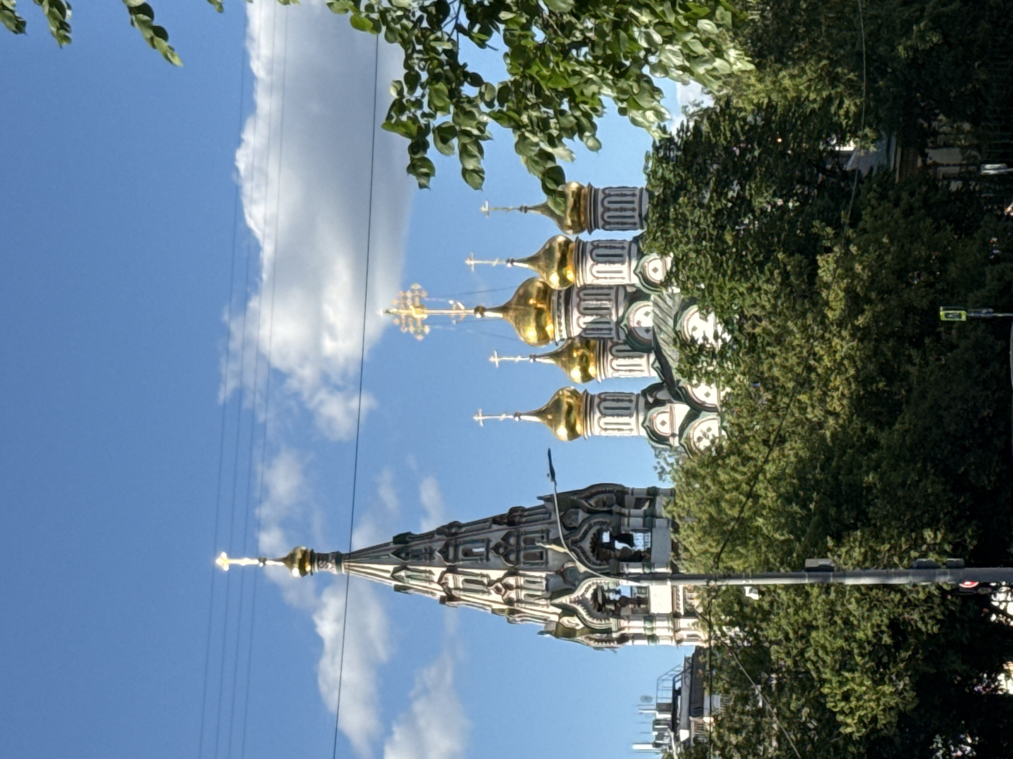 The image shows a clear blue sky with scattered white clouds, partially obscured by lush green tree branches in the foreground. A tall, ornate building with multiple golden domes and intricate white and green detailing rises against the sky. One prominent spire, also adorned with white and green architectural elements, dominates the lower portion of the image. The building appears to be a church or cathedral. Several golden crosses are visible atop the domes and spire. Power lines stretch across the sky. A traffic light and a street sign are visible in the lower right portion of the image. The sun is high in the sky, suggesting it is daytime. The overall impression is a bright, sunny day in an urban environment, likely Moscow, Russia, given the architectural style of the building. No people are visible, and no text is discernable.