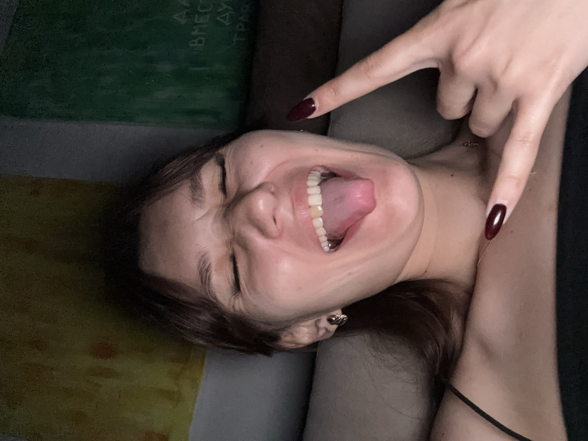 This is a close-up, low-angle selfie of a young woman with dark hair and closed eyes, sticking her tongue out. Her head is tilted to the right. Her tongue is a light pink and curled upwards. She has her eyes squeezed shut, and her eyebrows are furrowed. Her mouth is open, revealing her teeth. She is wearing a black top with a thin strap. A thin gold necklace is visible around her neck. Her right hand is raised, with her index and pinky fingers extended in a "rock on" gesture, while her other fingers are curled. Her fingernails are long and painted a dark burgundy color. Her left hand is also visible, with fingernails painted the same dark burgundy color. The background is out of focus but appears to be a couch or bed. There are some areas of dark green and yellow in the background, possibly indicating fabric or a wall. The lighting suggests it is likely nighttime or indoors. No specific location cues are visible in the immediate foreground.
There is some faint, smudged text visible in the upper left quadrant of the image, appearing to be in Cyrillic. The visible letters suggest words like "ВМЕСТ" and possibly "ТРАВ".