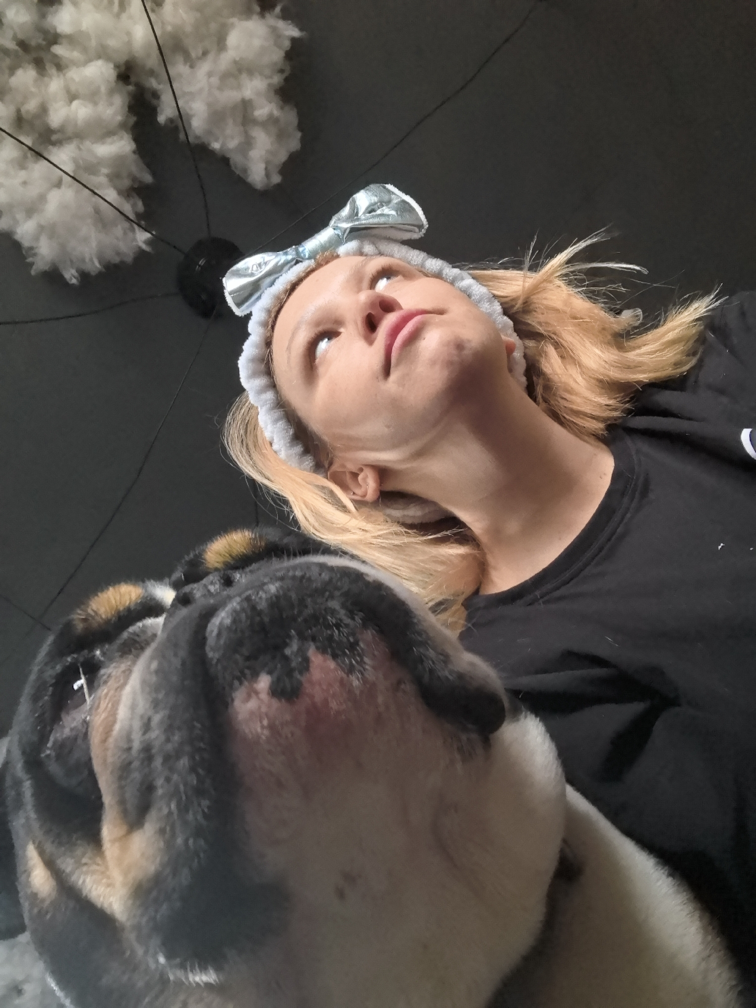 A low-angle shot shows a woman and a bulldog. The woman is wearing a light-colored headband with a bow and a black t-shirt. She is looking up and to the left. The bulldog's face fills the bottom left of the frame. Above the woman is a dark ceiling with a light fixture and cotton-like material hanging from it. The location is unknown, and there is no indication that the location is Noviy, Russia.