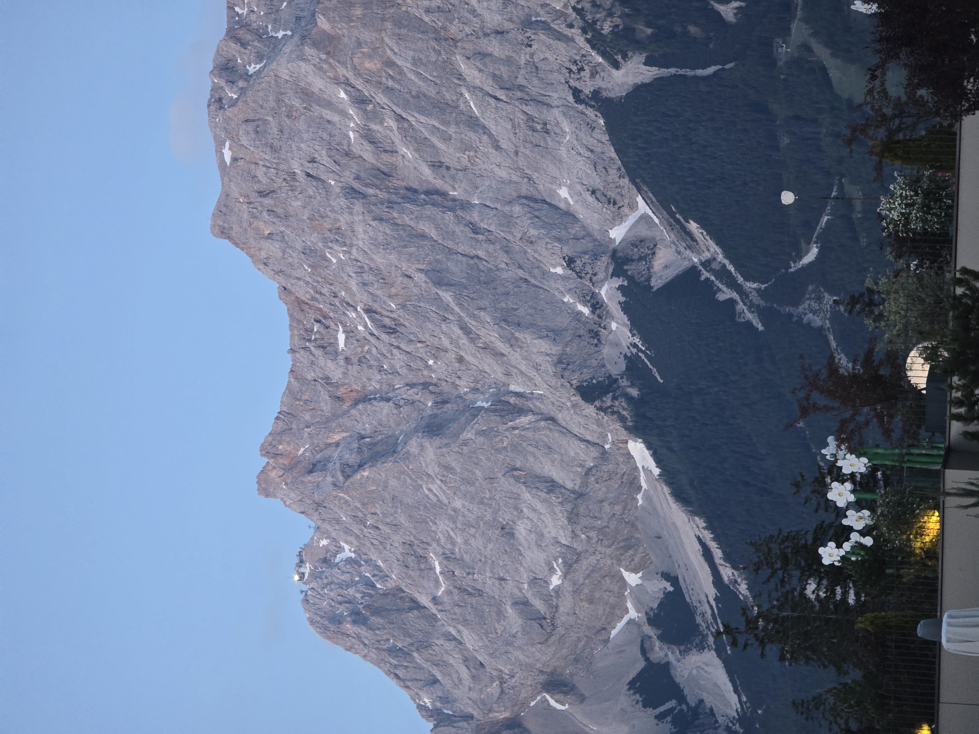 The image captures a grand mountain landscape at dusk or dawn, viewed from an elevated position, likely a balcony or terrace in Lermoos, Austria.
In the rotated view (where the sky is at the top), a massive, rugged mountain range dominates the left two-thirds of the frame. The mountain is predominantly grey rock with visible striations and patches of white snow on its slopes and ledges. A prominent peak on the far left of the mountain range features a distinct profile against the clear sky. Further down the mountain, towards the left, a small, illuminated structure, possibly a mountain station or observatory, is visible.
To the right of the mountain, a deep valley unfolds, densely covered in dark green coniferous forest, with some lighter grey, possibly rocky or scree-covered, areas visible within it.
In the far right foreground, elements of a man-made structure are visible. These include a dark railing, several white decorative flowers, and warm yellow lights emanating from what appears to be a building or balcony area.
The sky above the mountain and valley is a clear, light blue, devoid of clouds. The soft, indirect light on the landscape combined with the illuminated foreground suggests either sunrise or sunset. There is no visible text in the image.