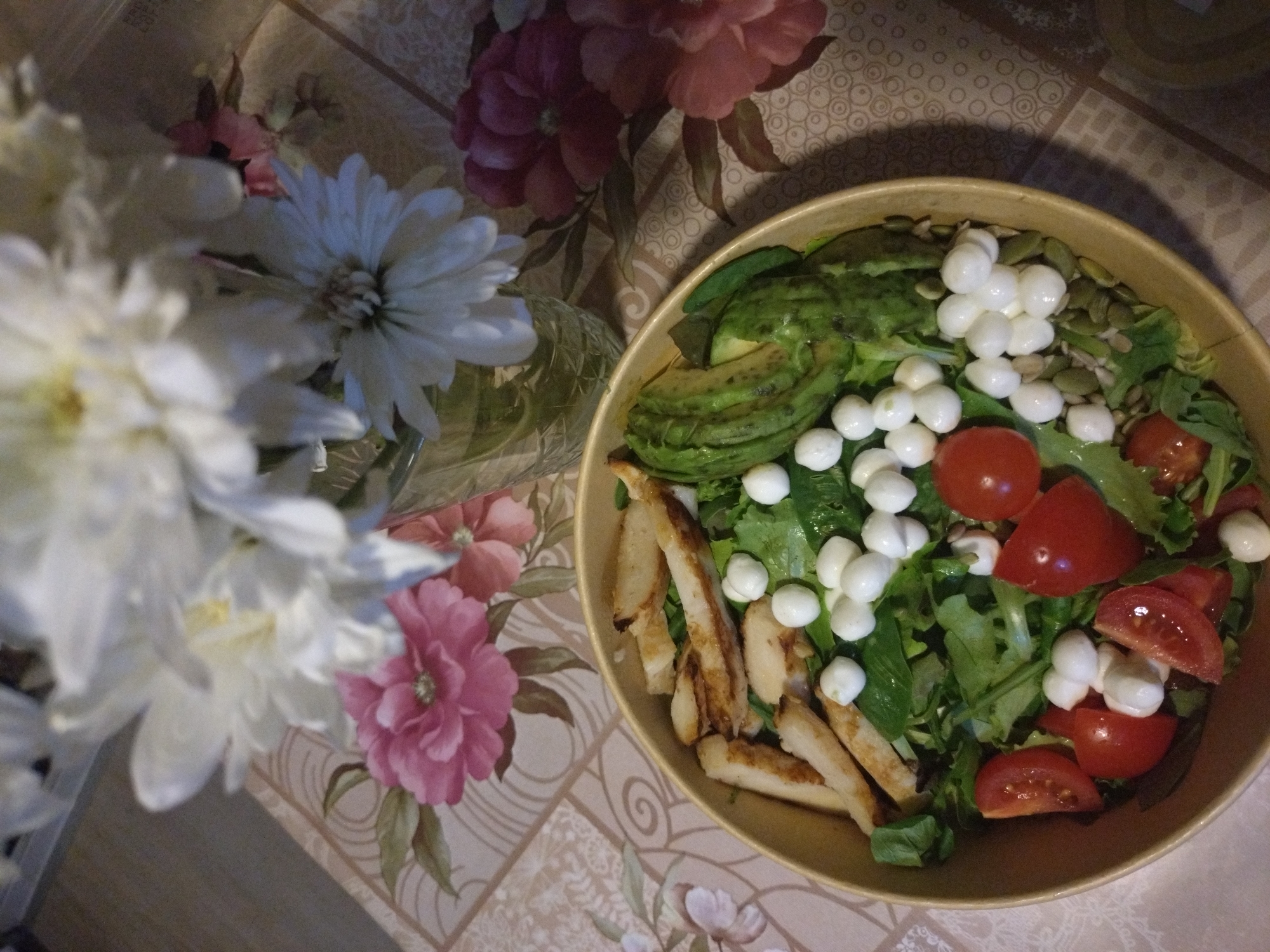 A top-down, eye-level shot shows a salad in a bowl next to a vase of flowers on a tablecloth. The salad contains avocado slices, small white cheese balls, sliced tomatoes, green leaves, grilled chicken strips, and green seeds. The flowers are white and pink. The tablecloth has a geometric and floral pattern. The location is presumed to be Stara Zagora, Bulgaria, but there's no specific visual element confirming this.