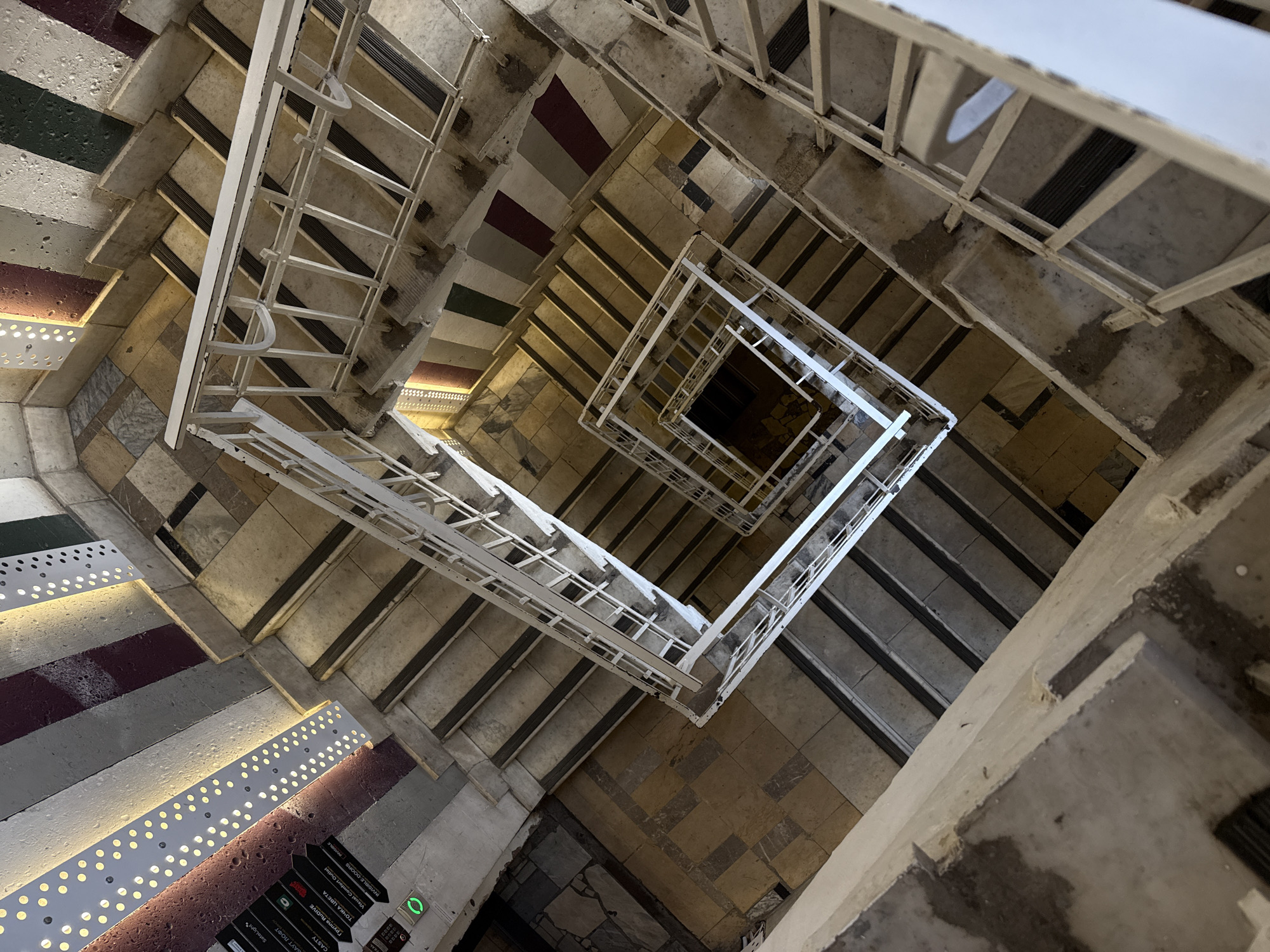 The image presents a bird's-eye view looking down a square, multi-story staircase inside a building, possibly a commercial or public space. The staircase features white metal railings and patterned stairs. Some stairs are illuminated with rows of small, round lights. The steps exhibit different materials and designs, including marble and colored stripes.
In the lower left corner, there are black signs with white text, some of which are readable as "BAUT LOFT", "CASTY", "Группа RuDIVE", "ТОЧКА ЦВЕТА", "Visual Comfort Outlet", and "INVISIBLE-DOORS". A green, circular emergency light is also visible near the signs.
Based on the Cyrillic text and signs, the location is Moscow, Russia. The presence of the signs indicates that the staircase may be part of a multi-tenant building or shopping complex.
