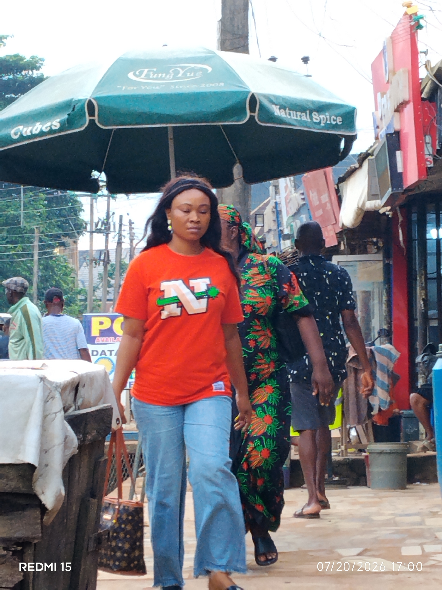 In Umuode, Nigeria, a woman walks forward on a sidewalk under a large green umbrella. She is wearing an orange t-shirt with an "N" logo and light blue jeans. She carries a patterned handbag. Behind her, other people walk along the street, including a woman in a brightly colored floral dress and a man in a black shirt. Shops and stalls line the street, with signs and merchandise visible. The scene appears to be a busy marketplace. Text on the umbrella reads "Fung Yue" and "Natural Spice." A timestamp indicates the date as July 20, 2026, at 17:00. The lighting suggests it is late afternoon.