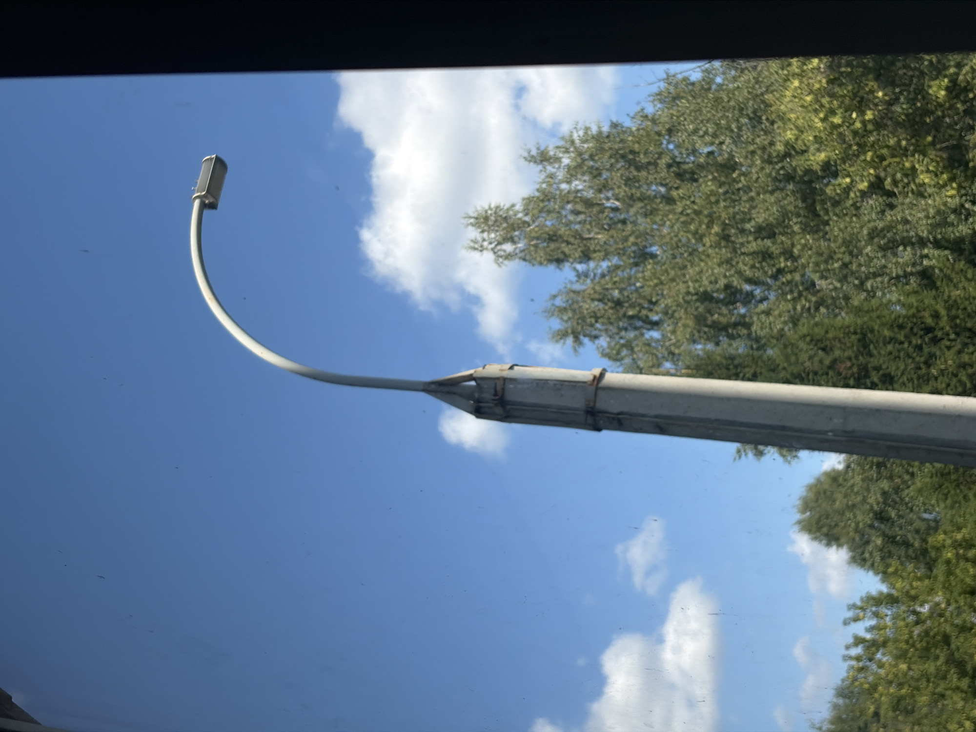 The image shows a street lamp against a bright blue sky with scattered white clouds. A portion of a concrete pole supports the curved arm of the street lamp, which terminates in a light fixture. Lush green trees are visible in the background, suggesting an outdoor, possibly suburban or park-like setting. The overall impression is a clear, sunny day. The image does not contain any information that would allow for specific identification of the location within Izhevsk, Russia.