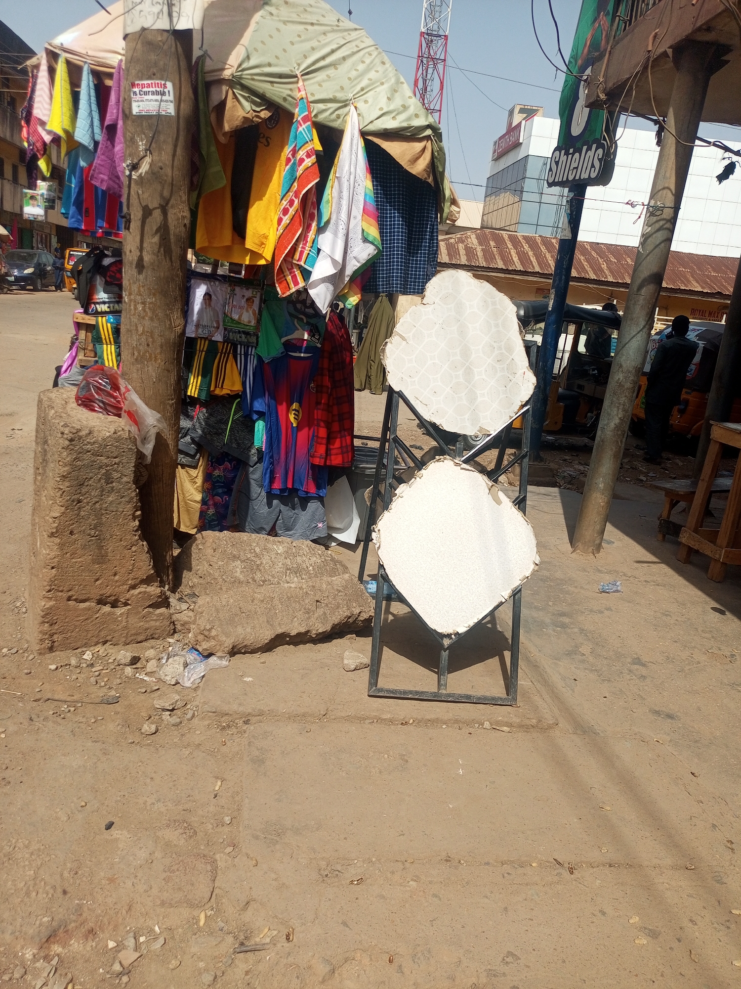 The image depicts a bustling street scene in Maiburiji, Nigeria, characterized by roadside commerce under clear, bright daylight, indicative of mid-day.
In the foreground and central to the composition is a makeshift stall built around a sturdy wooden pole. Various items of clothing and textiles, including colorful towels, t-shirts (some featuring sports team logos), and patterned fabrics, are displayed, hanging from ropes and shelves. At the base of the wooden pole are large, rough-hewn stone blocks, and some litter is scattered on the dusty ground. Attached to the wooden pole is a white sign with red text, prominently stating: "Hepatitis is Curable!". Below this, smaller, unreadable text is visible, possibly related to treatment or contact information. Further down the stall, another sign with "VICTORIA" and a logo is visible on a bag.
To the right of the stall, two white, somewhat octagonal or shield-shaped display boards are mounted on a black metal stand, facing the viewer. The upper board has a subtle geometric pattern, while the lower one is plain.
The mid-ground reveals typical street activity with vehicles and people. A dark-colored car is visible on the left, and a yellow tricycle (keke napep) is parked on the right. A person in a dark shirt stands near the tricycle, and another individual is partially visible further down the street.
The background features urban architecture. A modern building with white panels and reflective glass is prominent, clearly marked with "ZENITH BANK" in red lettering. Adjacent to it, a large green sign with the word "Shields" in white is visible. A tall red and white communications tower stands against the blue sky on the far left. The ground is a mix of dirt and worn asphalt, with shadows suggesting a high sun angle. A partially visible sign with "ROYAL MAXI" can also be seen in the background.