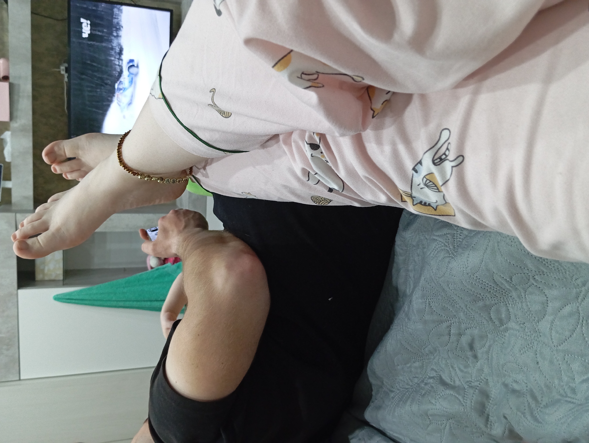A person in pink patterned pajamas is seated on a light blue patterned cushion. The person's bare feet are raised, with a gold ankle bracelet visible on one ankle. Another person is seen in a black shirt, holding a phone. A green towel is nearby. The background shows a television screen and white cabinetry. The location is unknown; there is no verifiable information confirming the city as Yurgamysh, Russia.