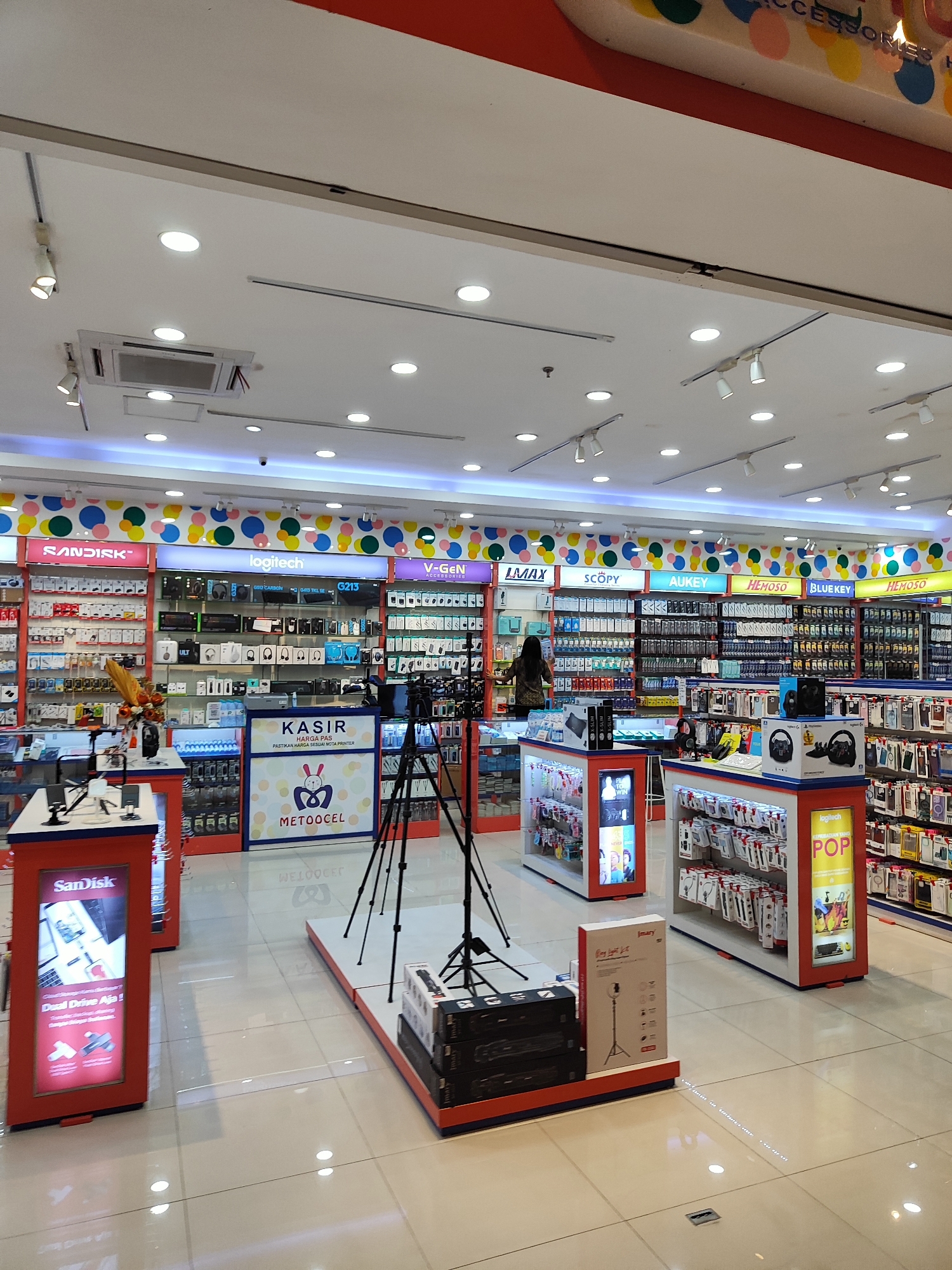 The image depicts the interior of a well-lit electronics store, likely located in Sempusari, Indonesia, as indicated by the store's branding and visible text. The store features extensive displays of consumer electronics, including memory cards, headphones, phone accessories, and cameras. The overall aesthetic is modern and organized, with brightly colored branding and clear product shelving.

In the foreground, a red display stand with a "Sandisk" advertisement is visible. To the right, a checkout counter with "KASIR" (cashier) signage and a "METOCEL" logo is present, indicating a point of sale. Several camera tripods are also set up in the central area of the store, suggesting a display or demonstration of camera equipment.

The shelves are stocked with a variety of electronic items, organized by brand and product type. Brands such as "Sandisk," "Logitech," "V-Gen," "LMAX," "SCOPY," "AUKEY," and "HEMOSO" are prominently displayed. The store is illuminated by a combination of ceiling lights and display lighting, creating a clean and professional ambiance.

A single female customer, dressed in a dark top, is visible near the "V-Gen" section, appearing to browse the merchandise. No other individuals are clearly discernible in the image. The flooring is a light-colored tile, reflecting the overhead lights. The ceiling features a modern design with recessed lighting and air conditioning units. The overall impression is that of a typical retail environment for electronics.