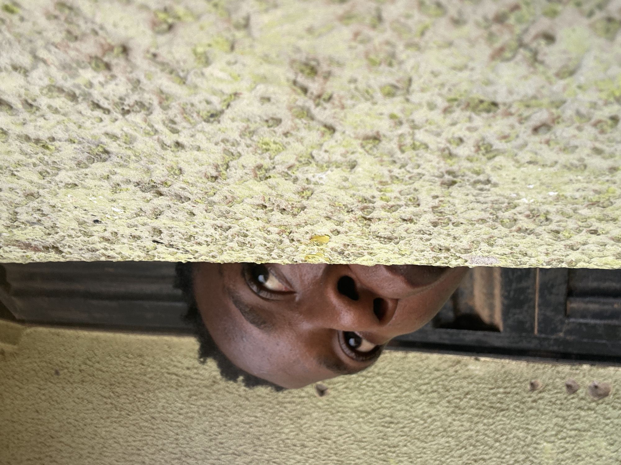 A person's face peers through a narrow gap between two rough, textured surfaces. The upper surface is a light, mottled color, while the lower surface is a solid, darker shade. The person's eyes are visible, looking upwards. The overall composition suggests a low-angle perspective, with the viewer positioned below the person looking through the gap. The location is in the city of Unknown, Unknown.