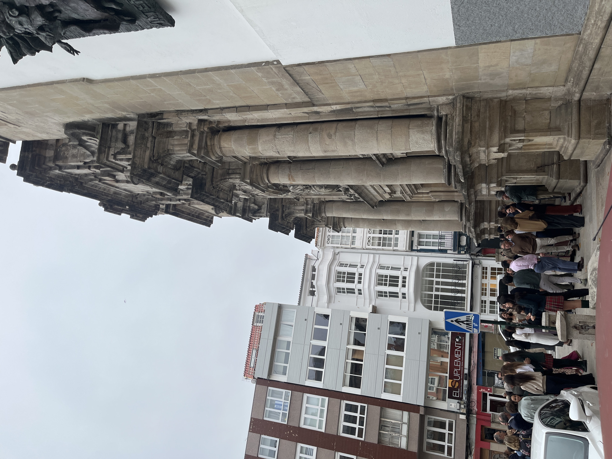 The image captures a street scene in A Coruña, Spain, featuring a section of the Iglesia de San Jorge, a baroque church. The church's facade is visible with its stone columns and detailed architectural elements. A group of people are lined up, possibly waiting to enter the church or a nearby building. A street sign with a pedestrian crossing symbol is visible, as well as storefronts with signs in Spanish. The buildings in the background have multiple windows and varying architectural styles. The sky is overcast.