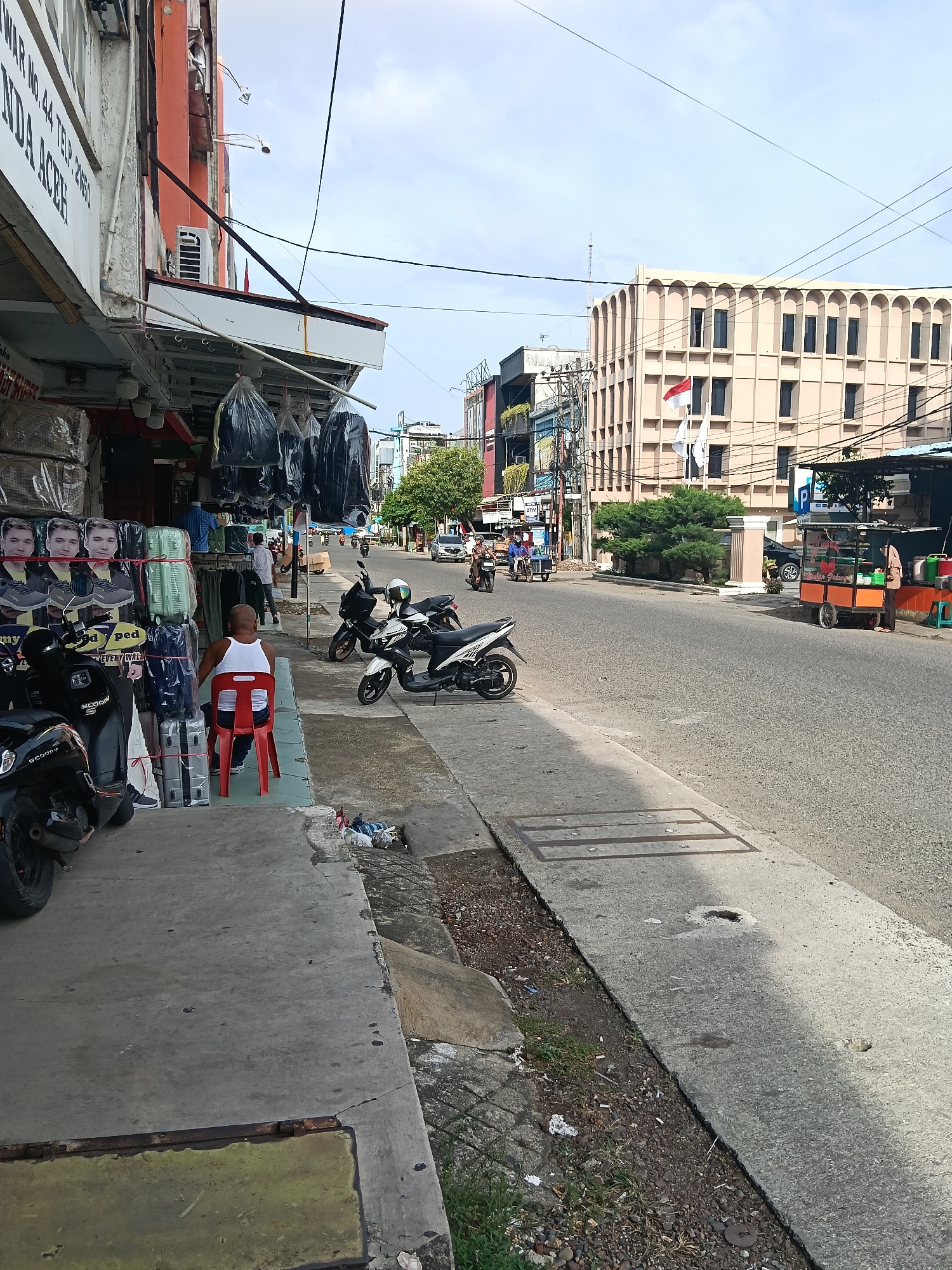 The image depicts a street scene in Banda Aceh, Indonesia. On the left, a storefront is visible, with merchandise displayed outside, including suitcases and posters of men. A person wearing a white tank top sits on a red chair in front of the store. Several motorcycles are parked along the sidewalk. The street itself is paved, with traffic including cars and motorcycles. In the background, a large, light-colored building with arched windows stands prominently. A food cart is situated on the side of the road. The sky is partly cloudy. The scene suggests an active commercial area on a daytime.