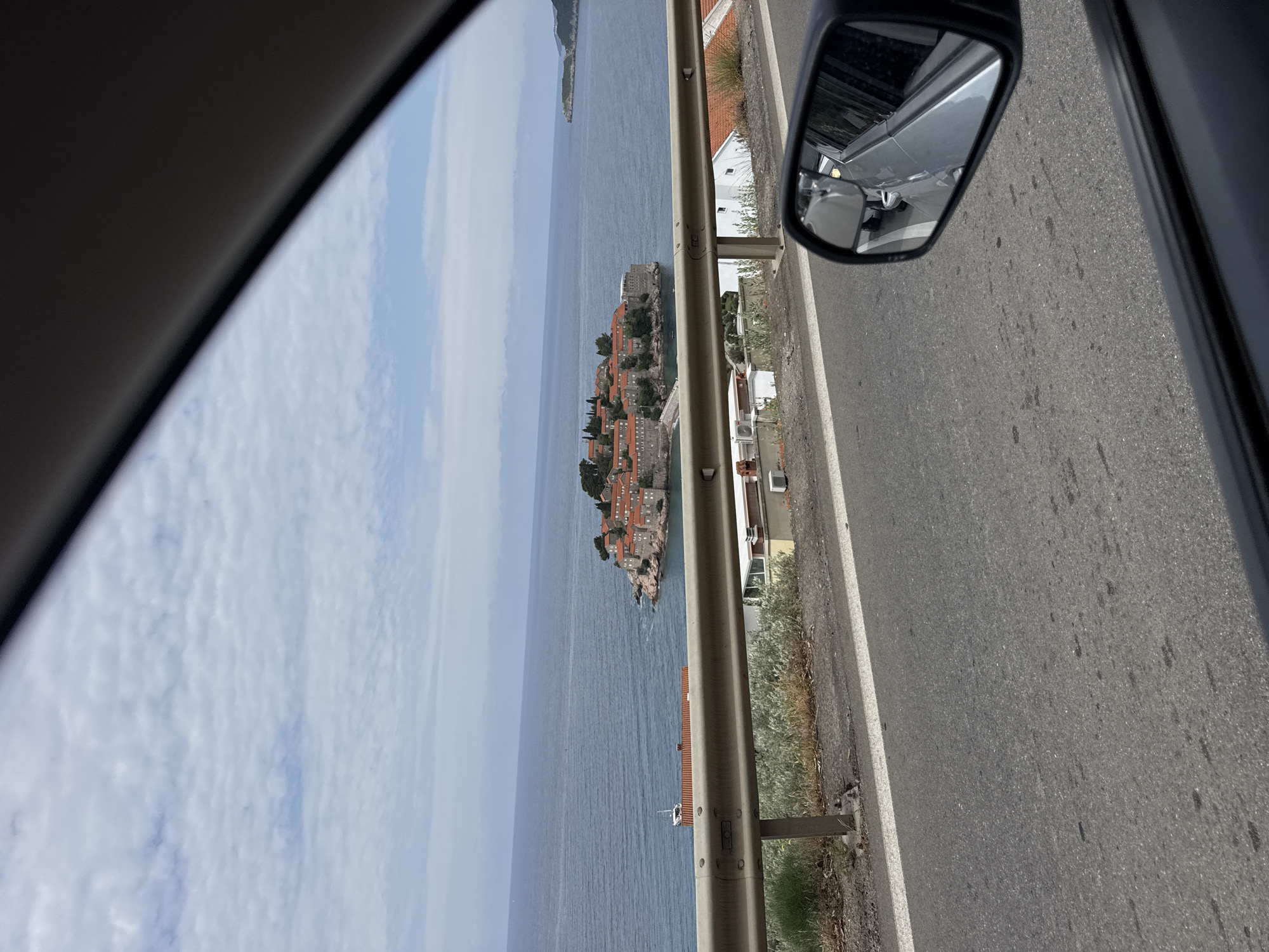 The image presents a picturesque coastal view captured from the interior of a vehicle, looking out through the side window. The photograph is oriented vertically, as if rotated 90 degrees clockwise from a landscape perspective.
In the foreground, the dark frame of the car's window is visible in the upper left, while on the right, a side rearview mirror reflects a portion of the vehicle's silver body and the adjacent asphalt road.
A sturdy gray metal guardrail spans horizontally across the middle section of the frame, acting as a boundary between the road and the steep incline to the sea. To the right of the guardrail is a section of an asphalt road with a clear white painted line along its edge. Beyond this, overgrown dry grass and sparse vegetation line the roadside, and parts of white-painted buildings with orange or red roofs are visible on the land side.
The main subject beyond the guardrail is the deep blue Adriatic Sea, stretching towards the horizon under a sky dotted with scattered white clouds. In the mid-distance, a prominent and distinctive small island or peninsula, identified as Sveti Stefan in Montenegro, emerges from the water. It is densely covered with numerous stone buildings, all featuring traditional reddish-orange tiled roofs, interspersed with green trees. A thin strip of land or causeway appears to connect the island to the mainland, although much of it is obscured. Another distant landmass or island can be faintly seen further out in the sea.
The scene depicts a bright, sunny daytime, suggesting pleasant weather conditions. The implied activity is traveling by car along a coastal road, offering a panoramic view of this iconic Montenegrin landmark. No discernible text is visible in the image.