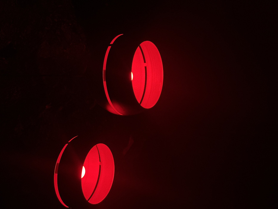 Two cylindrical, red-lit light fixtures illuminate a dark space. The fixtures are hollow, with a segment missing from their circumference. A bright, centralized point of light shines within each cylinder, casting an eerie glow. The top fixture is slightly tilted upwards, while the bottom one faces forward. The background is shrouded in darkness, enhancing the contrast and focusing attention on the glowing red forms.